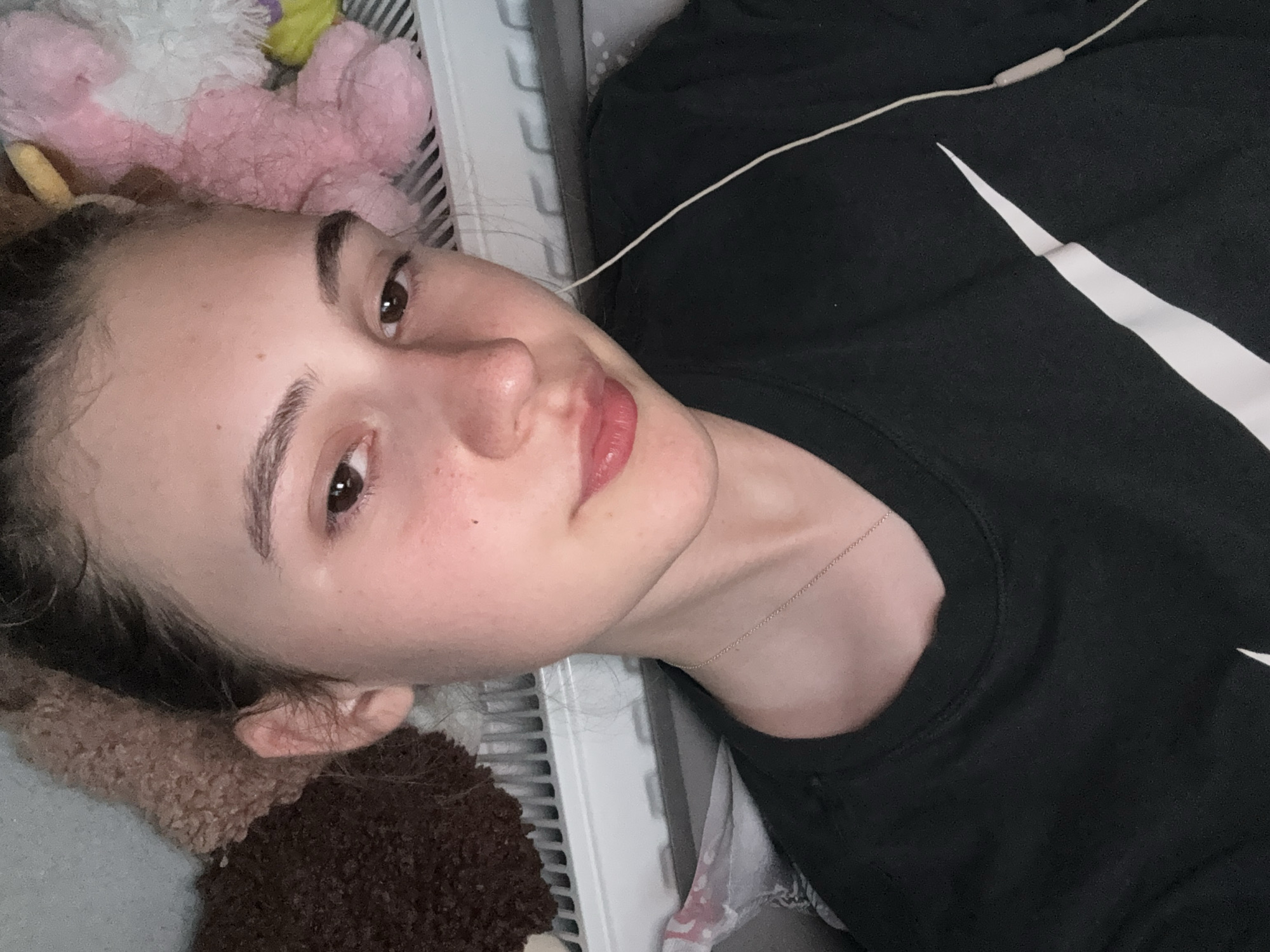The image shows a close-up, chest-up view of a young female. She has light skin with some freckles, dark brown eyes, and dark brown hair pulled back. Her lips are slightly parted and painted pink. She is wearing a black t-shirt with a white swoosh logo on the left chest. A thin silver necklace is visible around her neck. White earbuds are in her ears, with the wire running down her t-shirt. In the background, there are plush toys, a radiator, and a patterned fabric. The lighting is dim. The scene is indoors. There is no information in the image to determine the location in the city of Panagyurishte, Bulgaria.