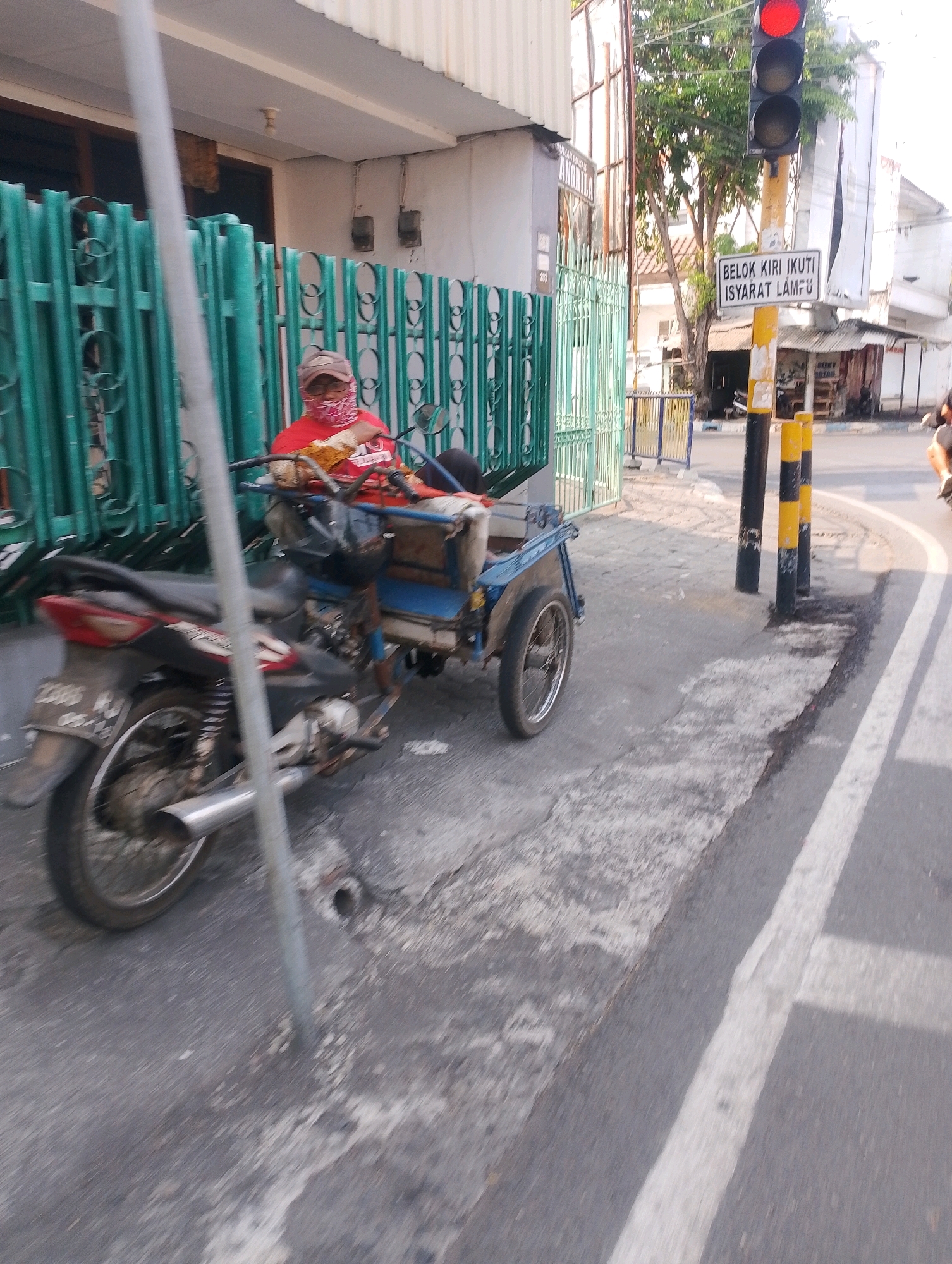 The image shows a street scene in Probolinggo, Indonesia. In the foreground, a three-wheeled motorcycle, commonly known as a *becak motor* or *bentor*, is parked on the side of the road. The motorcycle has a sidecar attached, designed to carry passengers or goods. The driver, wearing a red shirt and a patterned face covering, is seated on the motorcycle. The *becak motor* has a license plate that reads "AE 2385 X".
Behind the *becak motor*, a green metal fence is visible, separating the road from a building. Further in the background, a traffic light is illuminated with a red signal, indicating vehicles should stop. A street sign below the traffic light reads "BELOK KIRI IKUT ISYARAT LAMPU" which translates to "TURN LEFT FOLLOW TRAFFIC SIGNAL". The environment appears to be a typical urban street with buildings, trees, and a paved road. The lighting suggests it is daytime.