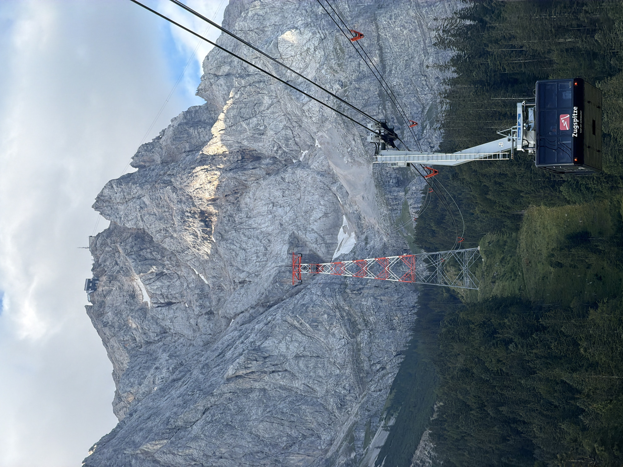 A gondola with "Zugspitze TOP OF GERMANY" written on it hangs in the foreground, suspended by cables. Behind it, a tall, red and white lattice tower stands on a grassy slope dotted with evergreen trees. Further in the background, a large, craggy, grey mountain dominates the scene, with snow patches visible on its upper reaches. A building and a smaller tower are visible on the summit of the mountain. The sky above is partly cloudy. This is the Zugspitze, Germany's highest peak, located near Grainau.