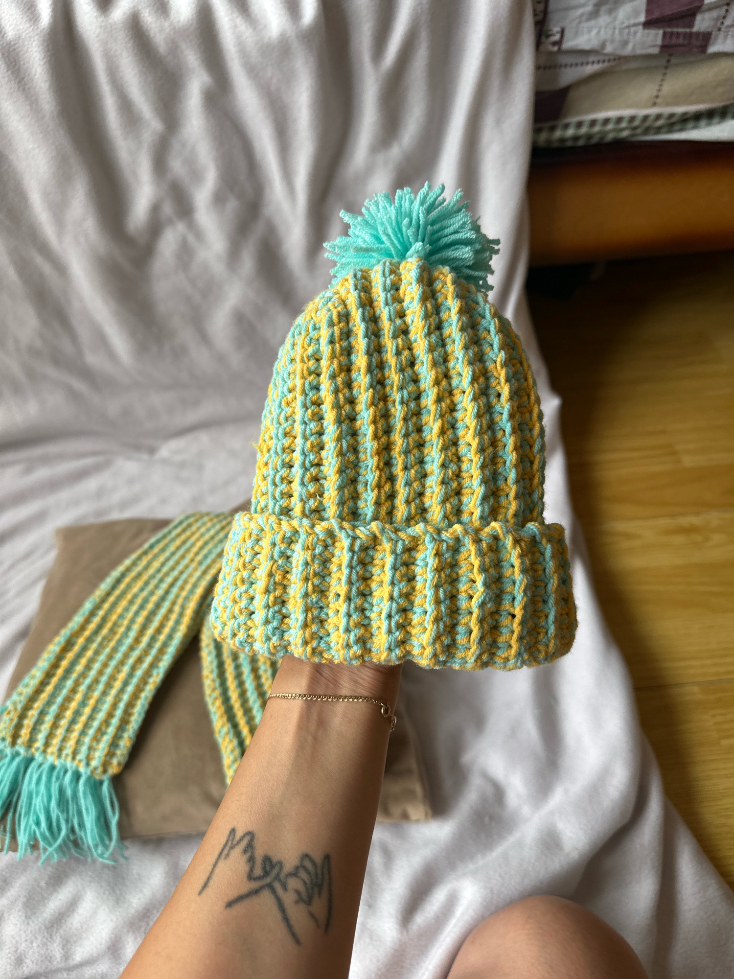 A person's arm, adorned with a thin bracelet and a simple tattoo of two hands making a pinky promise, holds a knitted hat and scarf set. The set is primarily yellow and turquoise, with the hat featuring a turquoise pom-pom on top. The items are placed against a white, textured surface, likely a blanket or sheet. A brown object, possibly a pillow, is partially visible behind the scarf. The background shows a wooden surface and a stack of fabric. This image does not provide any information about the city of Gyumri, Armenia.