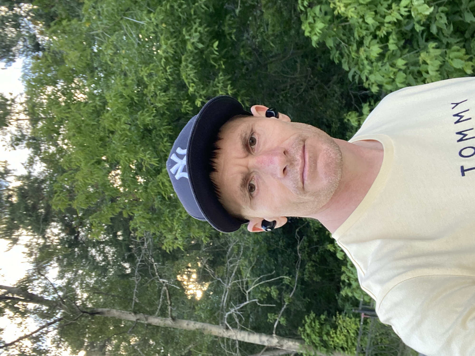 A close-up, selfie-style shot shows a fair-skinned man wearing a light yellow t-shirt with "TOMMY" written on it in dark blue letters, a dark blue baseball cap with a white "NY" logo, and black wireless earbuds. He has short, dark brown hair and is looking directly at the camera with a neutral expression. The background consists of lush green trees and foliage, suggesting an outdoor, possibly wooded, setting. Sunlight filters through the trees, creating a slightly dappled effect. There are no visible landmarks or architectural elements that would indicate a specific location within Yekaterinburg, Russia. The image appears to be taken during daylight hours.