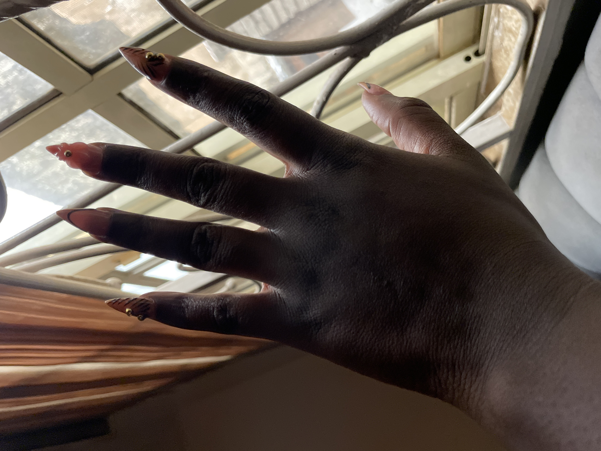 A hand with long, elaborately decorated acrylic nails is held up against a window with metal bars. The nails are painted with a brown and black tiger stripe pattern and adorned with small gold beads and what appear to be small gold charms. The hand itself is dark-skinned. Behind the hand, the window shows a blurred view of what appears to be buildings or structures outside, suggesting an urban or semi-urban environment. The lighting in the image is dim, with light filtering through the window from an exterior source. The curtains behind the hand are a warm, muted brown color. This image was taken inside a residence in Keffi, Nigeria.