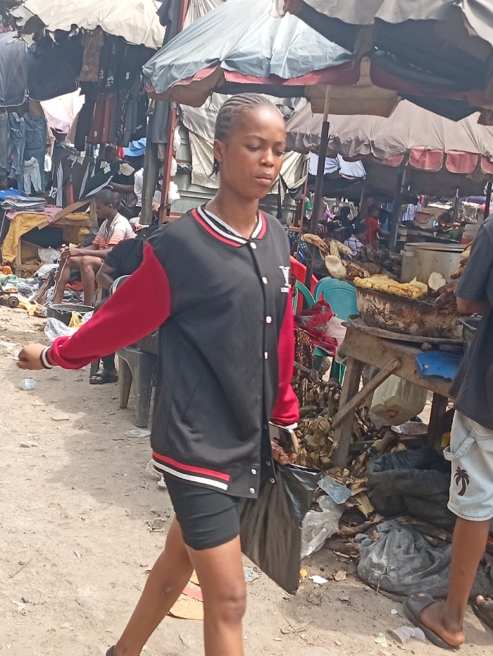 An outdoor market scene in Fegge, Nigeria, depicts a young adult female walking from left to right in the foreground. She has braided dark hair and wears a black bomber jacket with red sleeves, a white and red striped collar and cuffs, and a white 'Y' logo on the left chest, paired with black shorts. She carries a black plastic bag in her left hand, partially obscured. The unpaved dirt ground beneath her is littered with plastic refuse and organic debris, including plantain peels.
In the midground and background, multiple market stalls operate under large, light-colored tarpaulin awnings. The left side of the frame shows clothing stalls with various garments hanging from display racks, and an individual is seated on a low structure nearby. The right side features a food vendor's stall, where a wooden table supports cooking apparatus and prepared food items, including what appear to be grilled plantains or yams. A person is partially visible on the far right, standing near this food stall, wearing light-colored shorts and sandals. The scene is brightly illuminated by daylight, indicating active commerce.