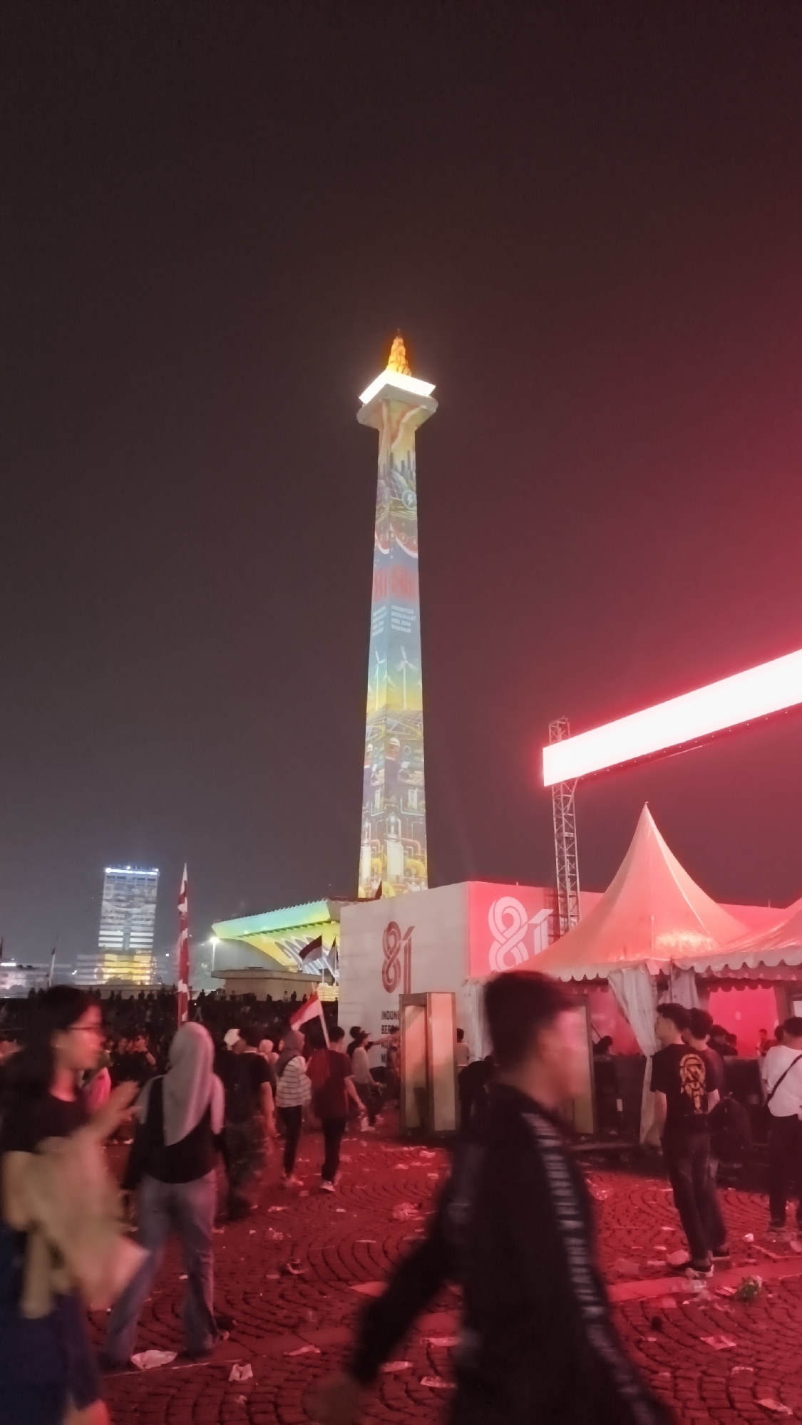The image depicts the National Monument (Monas) in Central Jakarta, Indonesia, at night. The monument is illuminated with projected colorful images and a flame at its apex. In the foreground, a crowd of people are gathered on a red, paved surface strewn with litter. Several tents, one brightly lit with red light, are visible in the middle ground. Indonesian flags are displayed on poles near the base of the monument. In the background, a tall, modern building stands against the dark sky.