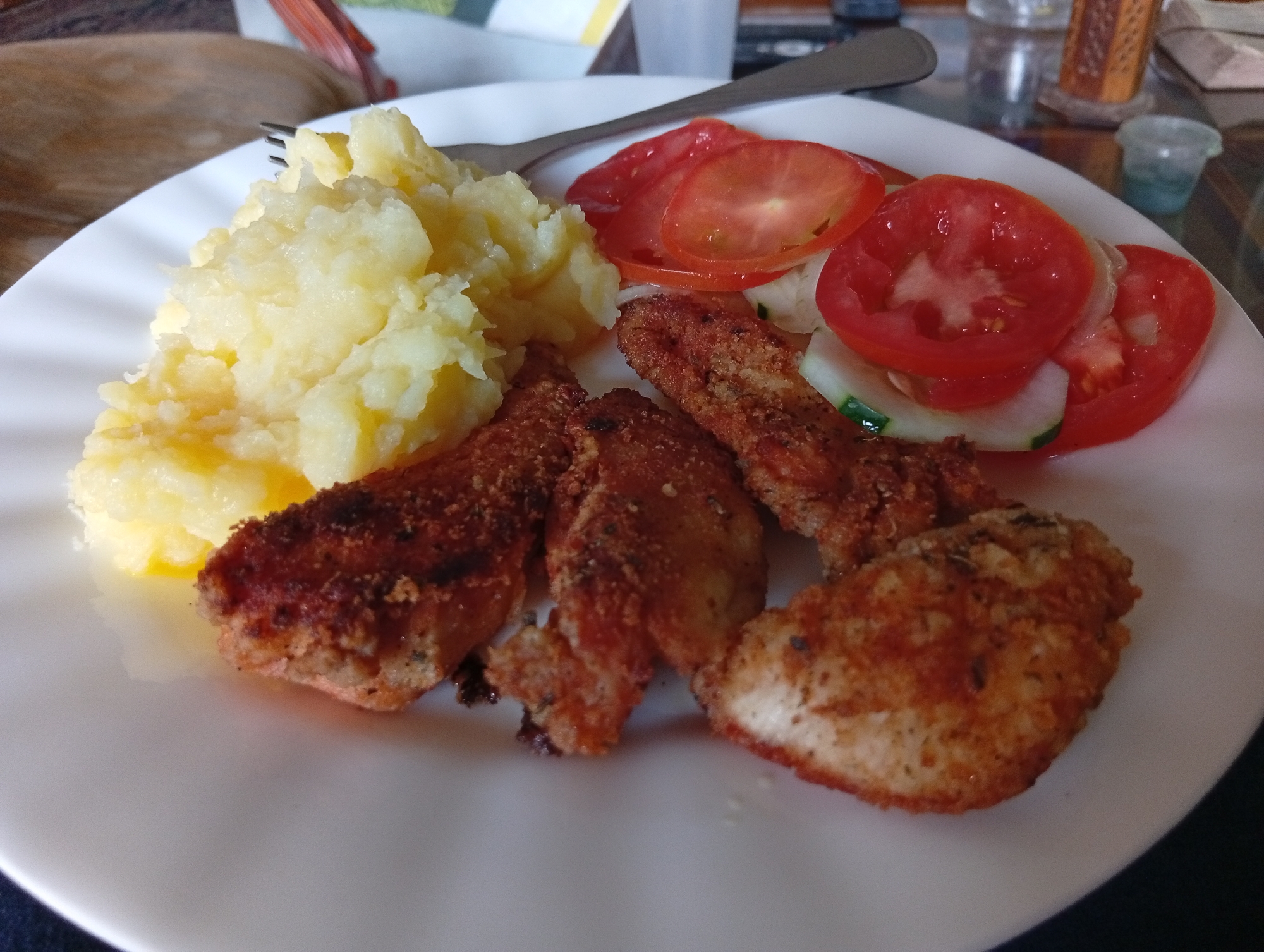 A plate of food, including mashed potatoes, fried chicken strips, and a tomato and cucumber salad, is displayed on a white plate. A fork rests on the plate, partially obscured by the mashed potatoes. The plate is situated on a glass-topped table. In the background, a wooden table or chair is visible, along with out-of-focus elements like a glass of liquid and a small container. The scene appears to be an indoor setting, possibly a dining area. The specific location in San Antonio de Los Altos, Venezuela, is not discernible from the image itself; this information would need to be provided externally.