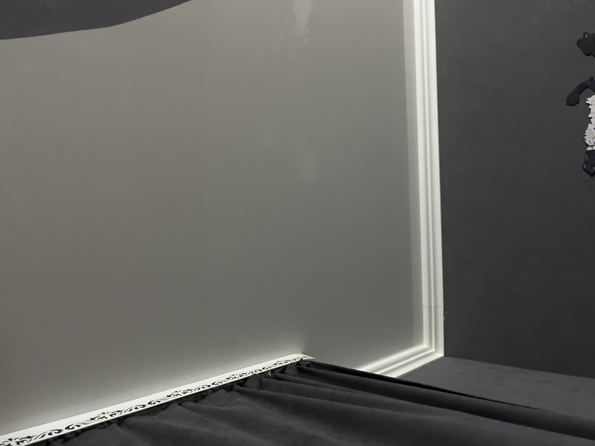 A close-up, low-angle interior shot shows a ceiling with a decorative cornice along the top of a dark gray wall. To the left, a dark gray curtain hangs down, partially obscuring the wall and cornice. The ceiling is a light, neutral gray, with subtle variations in lighting. There are no people or active events depicted in the image. The scene is static and appears to be a part of a room's interior decoration. The location is identified as Saratov, Russia.