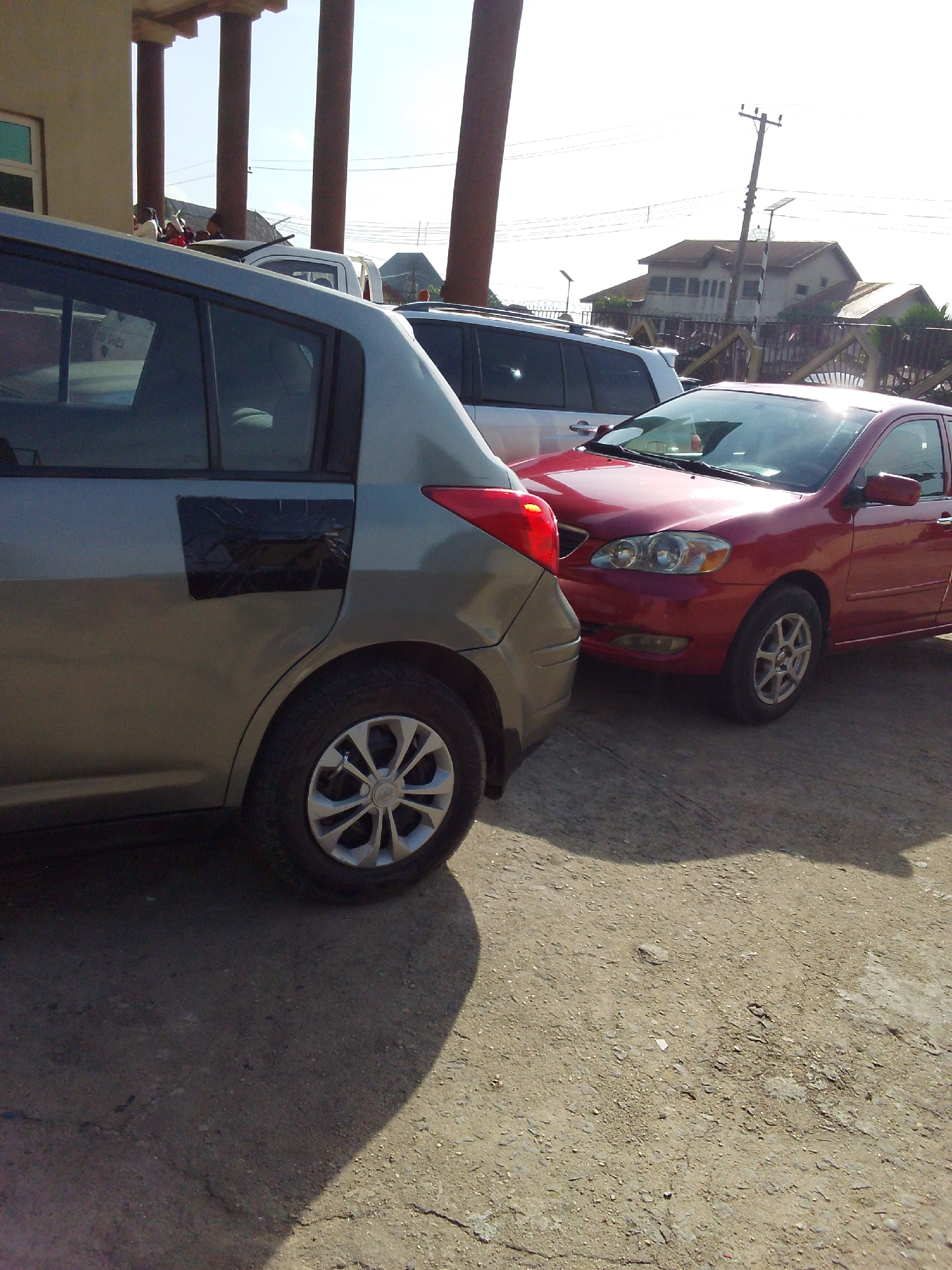 The image shows three parked cars on a dirt and gravel surface. The closest car to the left is a gray hatchback with a dark rectangle covering a portion of the rear passenger door. Next to it is a red sedan. Behind these two is a white SUV. In the background are several buildings and a utility pole. People appear to be gathered on the roof of one of the buildings. The location is Umuode, Nigeria.