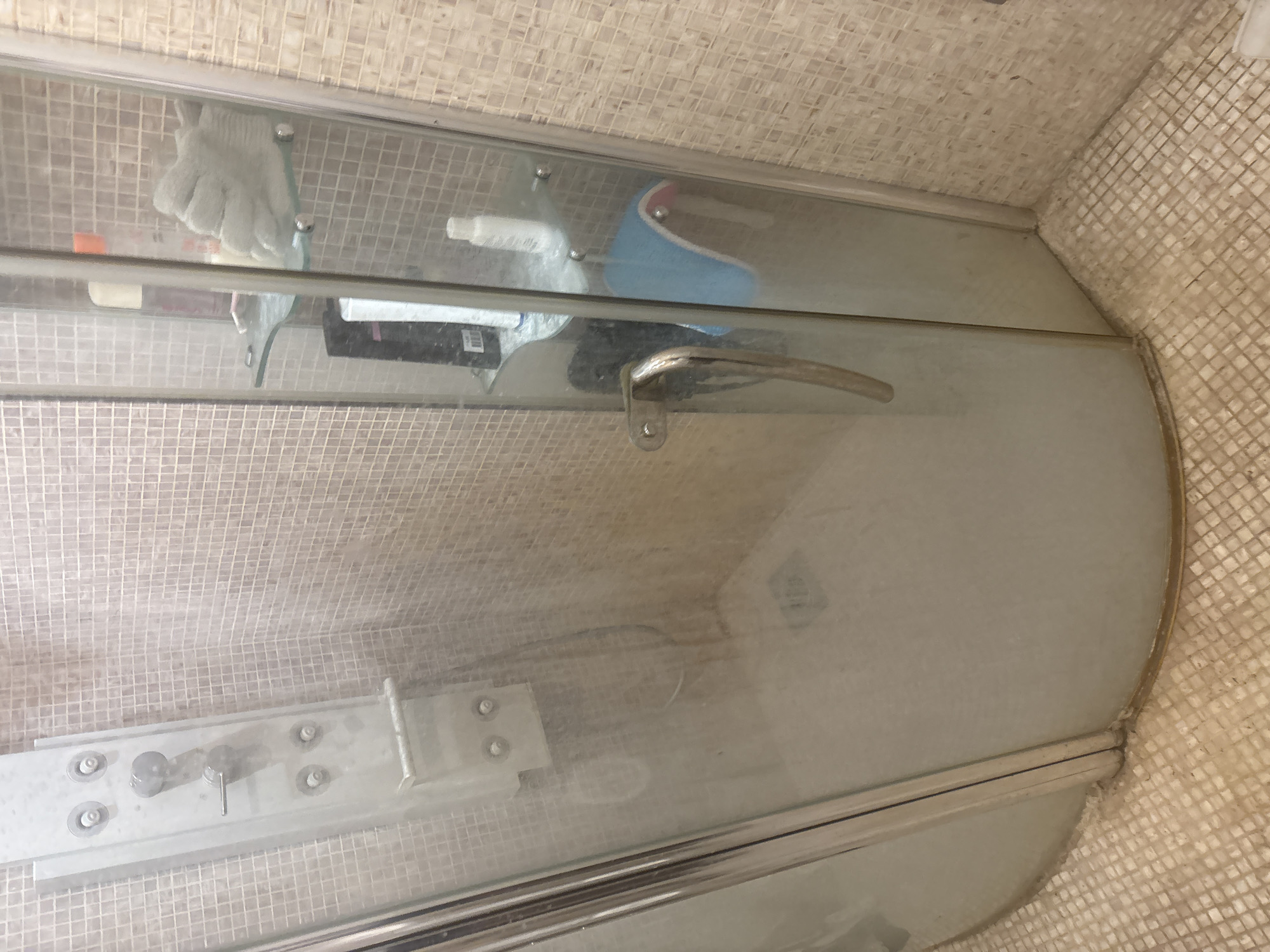 The image shows the interior of a shower stall with a glass door and tiled walls and floor. The shower stall is equipped with a multi-function shower panel. On a glass shelf inside the shower, there are several bottles of toiletries and a light-colored bath glove. The floor of the shower stall appears to be a drain. The overall impression is that of a functional, albeit somewhat dated, bathroom. The provided location of Orekhovka, Russia, is noted for context, but the image itself does not contain any specific geographical indicators to confirm this.