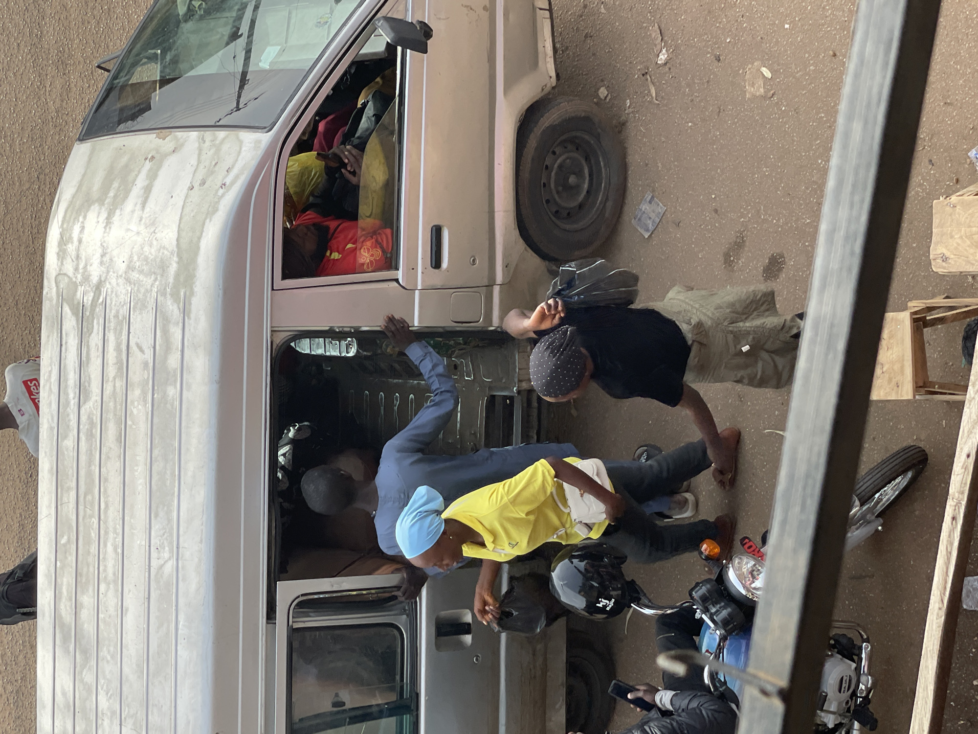 The image shows a group of people interacting with a light-colored van and a motorcycle on a dusty, outdoor surface. The van appears to be open, with several individuals inside and around it. One person in the van is wearing a red garment, and another is partially visible wearing a yellow shirt and a light blue headscarf. A person in a blue shirt is reaching into the van.
Outside the van, two men are standing. One, in a black shirt and patterned head covering, is partially obscured. The other man, wearing a dark shirt and patterned head covering, is looking downwards. A black motorcycle with a visible license plate is parked next to the van. The motorcycle has a red and clear headlight, and what appears to be a black helmet resting on its seat.
The scene suggests a moment of loading or unloading of goods or passengers, or possibly a repair or informal transaction involving the van and motorcycle. The ground is rough and uneven, suggesting a rural or informal urban setting. There is no clear indication of the time of day or weather, but the lighting seems natural, implying daytime.
One small piece of text is visible on a white bag in the upper right corner of the image, but it is illegible. There is also text on the helmet which reads "Hi". The overall mood appears utilitarian and busy, with people engaged in practical activities.