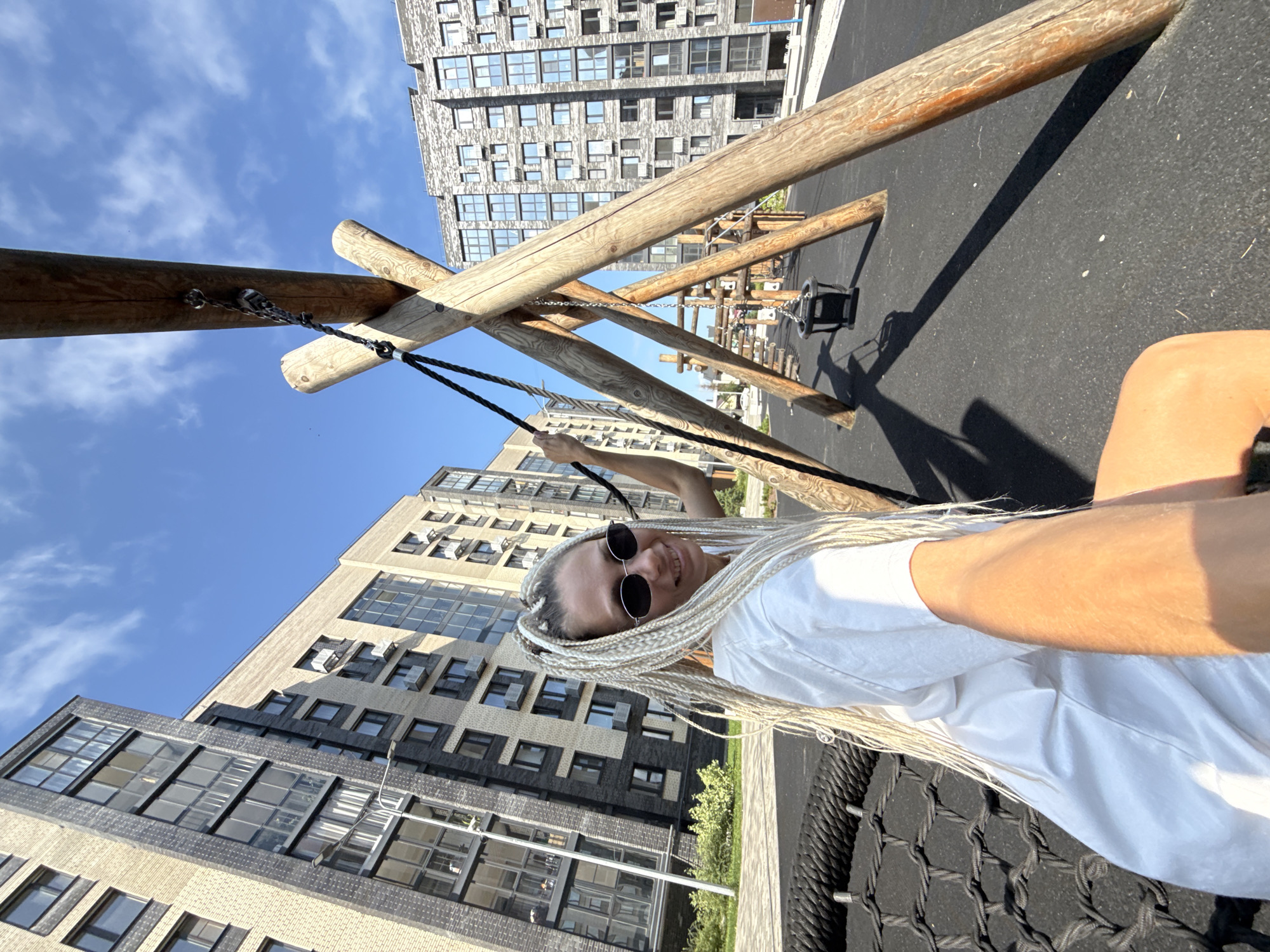 In Rusanovo, Russia, on a sunny day, a young woman with blonde braided hair and sunglasses is seated on a black net swing. She is wearing a white t-shirt. The swing set, made of large wooden logs, is located on a black rubberized playground surface. In the background, modern residential buildings with cream and dark gray facades rise under a bright blue sky with scattered white clouds. A child's swing bucket seat hangs from a chain within the A-frame structure of the wooden swing set.