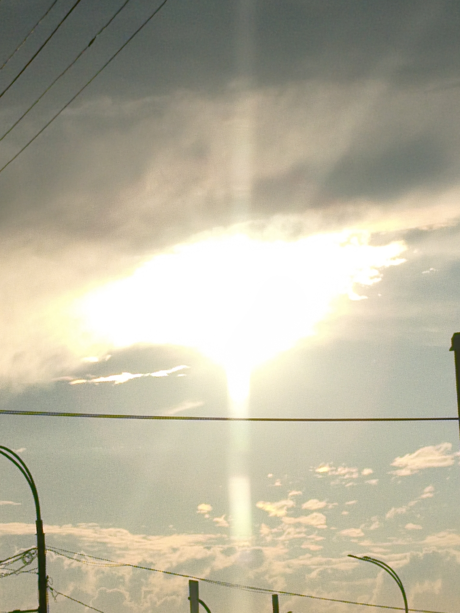 The image captures a bright sky with scattered clouds, illuminated by a strong source of light, possibly the sun, which appears to be behind a cloud formation and creating a bright, somewhat diffuse glow. The sky is a pale blue with varying shades of gray and yellow, indicating either sunrise or sunset, or perhaps overcast conditions with a break in the clouds.
In the foreground, several utility wires are visible, stretched horizontally across the frame. These wires are supported by poles and appear to be part of the city's infrastructure. On the left side, a dark pole with curved elements, likely a streetlight or part of a traffic signal, is partially visible. On the right side, another utility pole is also present. The ground is not visible in this image.
The scene appears to be an outdoor urban or semi-urban environment. The presence of overhead wires and utility poles suggests a developed area. While no specific landmarks are identifiable, the overall impression is that of a city skyline or a street view with infrastructure typical of Kemerovo, Russia. The image is characterized by its bright illumination and the contrast between the sky and the linear elements of the utility lines.
