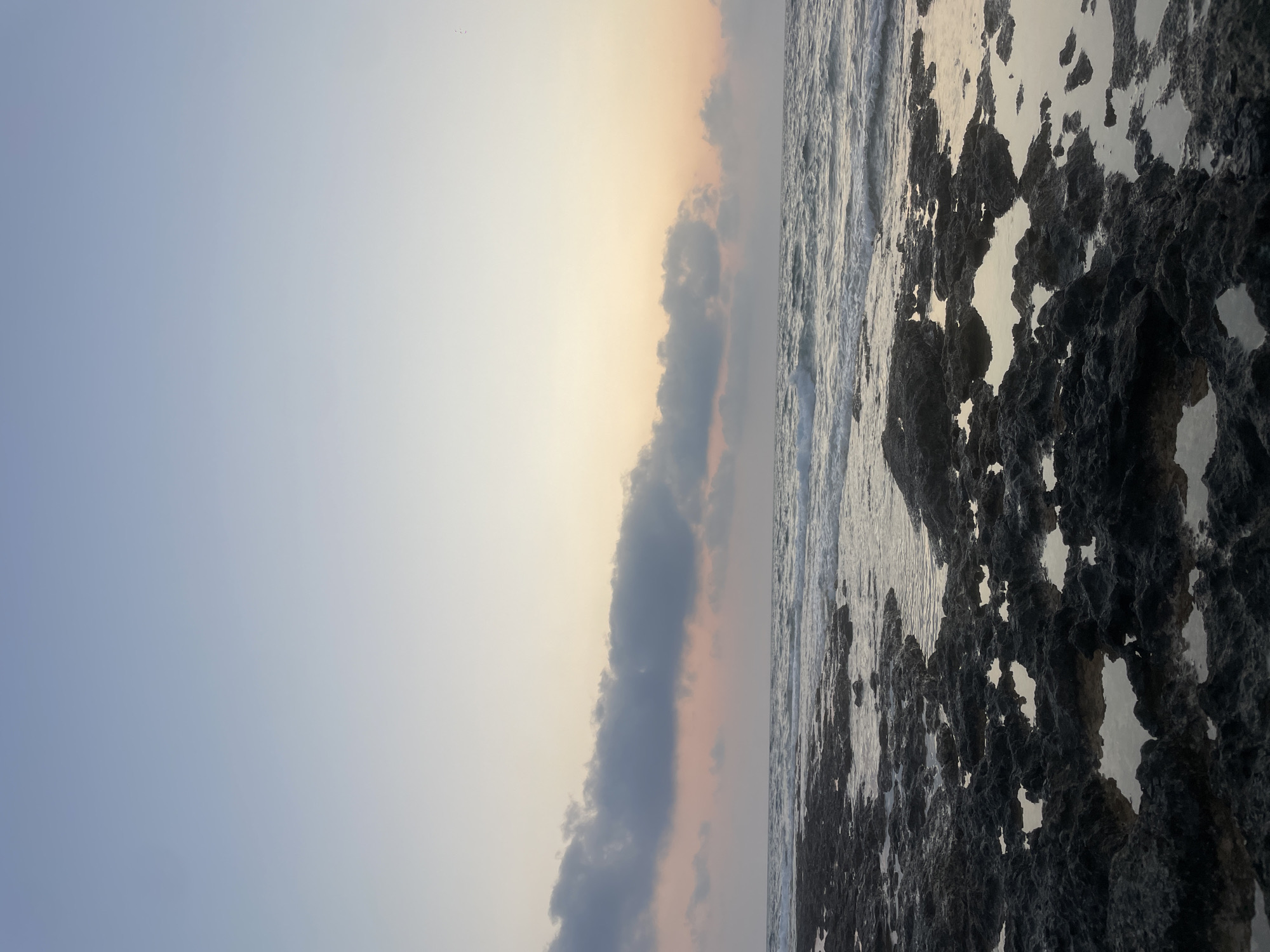 A serene seascape captures a rocky shoreline at dusk. The foreground is dominated by dark, jagged rocks, some of which are submerged in shallow pools of water that reflect the soft light of the fading day. Beyond the rocks, gentle waves roll in from the sea, creating a textured surface of white foam and shimmering water. In the distance, a line of clouds stretches across the horizon, tinged with the subtle hues of pink and orange from the setting sun, which is not directly visible but implied by the sky's gradient. The upper portion of the image displays a clear, pale blue sky that transitions into softer, lighter tones near the horizon. The scene depicts a natural, undisturbed coastal environment. Given the rocky coastline and the appearance of the sea, this location could be characteristic of many coastal areas within the Paphos Municipality, Cyprus, such as the Akamas Peninsula or areas near Petra tou Romiou.
