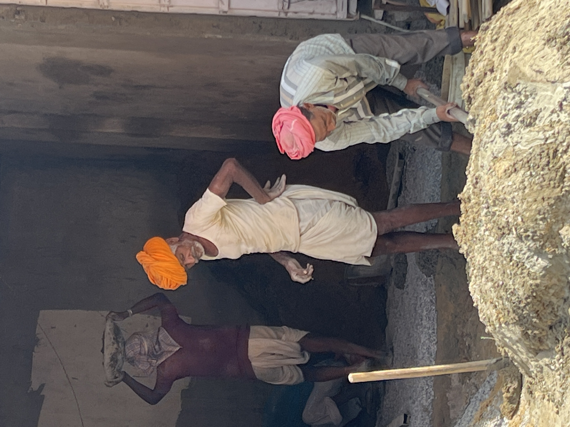The image depicts three men working at a construction site in Pushkar, India. In the foreground, a man wearing a pink turban and a striped shirt is using a shovel to mix materials in a large pile of what appears to be sand and cement. To his left, another man with an orange turban and a white shirt is standing and looking down, holding a wet cloth in his right hand. Behind them, a third man, his face obscured by a white cloth and wearing a maroon shirt and white dhoti, is carrying a basket filled with materials on his head. The environment appears to be an unfinished building with rough, textured walls and a gravel floor. A wooden plank or pole is visible in the left foreground, propped up against the pile of mixed materials. The lighting suggests it is daytime, possibly mid-morning or afternoon, casting shadows within the structure.