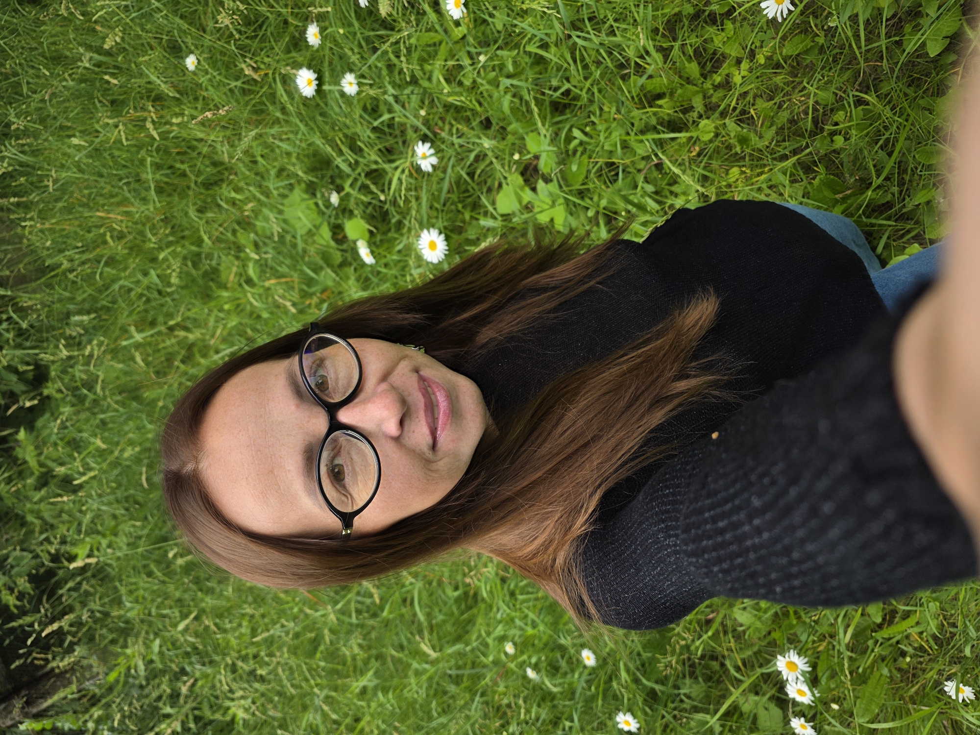 The media file is a close-up, overhead shot of a woman lying on green grass dotted with small white daisies. The woman, who appears to be in her late 20s or early 30s, is wearing black-rimmed glasses and a black knitted sweater. Her brown hair is spread out on the grass. The camera, presumably held by the woman herself, is capturing a selfie-style shot. The image is oriented vertically, with the woman's face at the top and her body extending downwards into the grassy field. The lighting suggests it is daytime, possibly late afternoon, given the soft light and lack of harsh shadows. The setting is a natural outdoor environment, likely a park or meadow, given the abundance of grass and daisies. The woman's expression is neutral, with her eyes open and looking towards the camera. There is no visible text in the image. The overall impression is one of peaceful relaxation amidst nature.