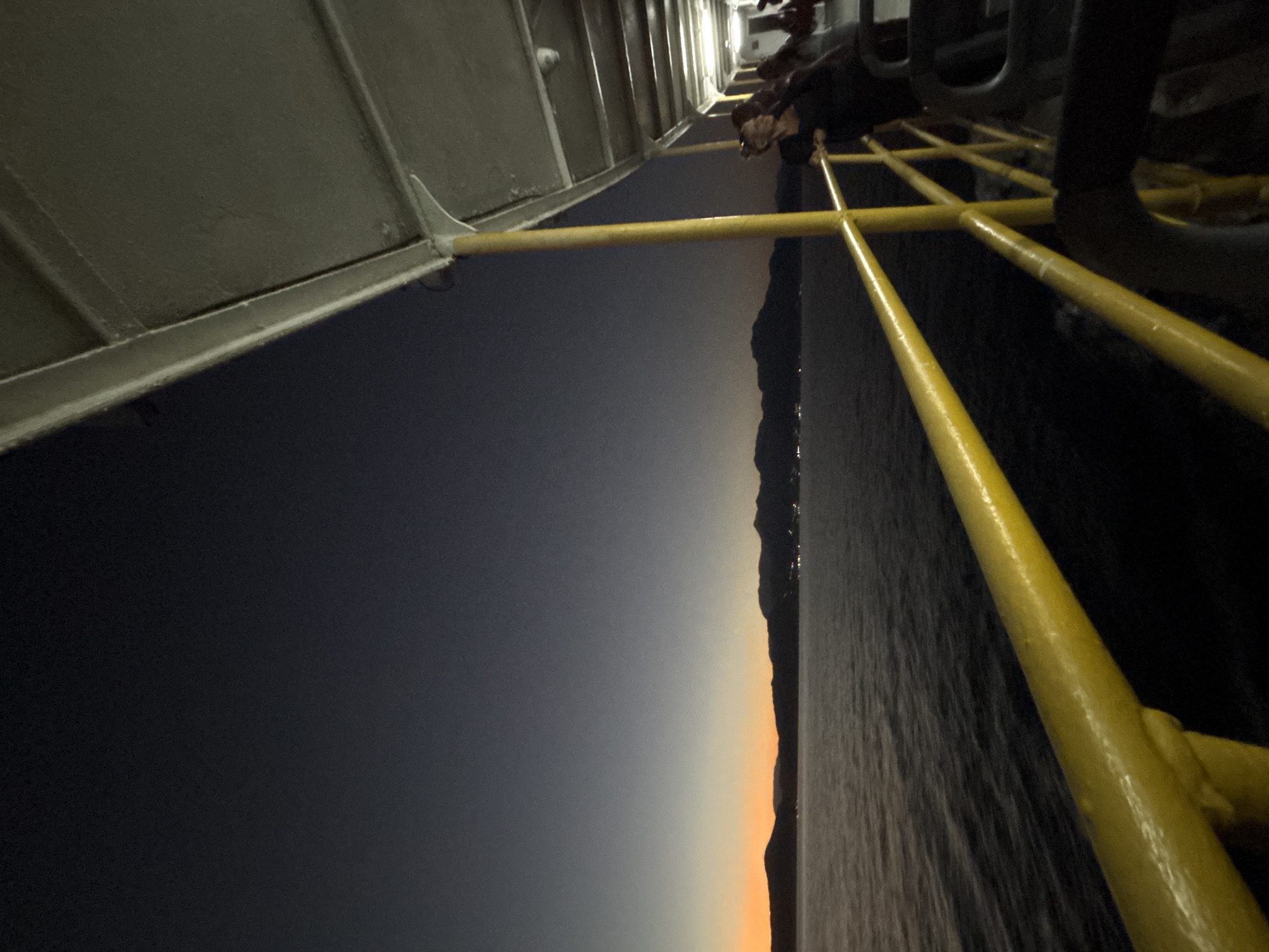 The image depicts a view from the deck of a ferry at dusk, looking out towards a coastline. The sky transitions from a deep blue at the top to a soft orange near the horizon, suggesting the sun has recently set. In the distance, a silhouetted mountain range borders the sea, with faint lights indicating human settlements. The ferry's yellow railing and structural elements are prominently visible in the foreground, suggesting the vantage point is from the upper deck. A few people are standing near the railing, looking out at the view. The water is calm, with gentle ripples reflecting the fading light. The scene suggests a tranquil evening journey. The location is the city of Angra dos Reis, Brazil.