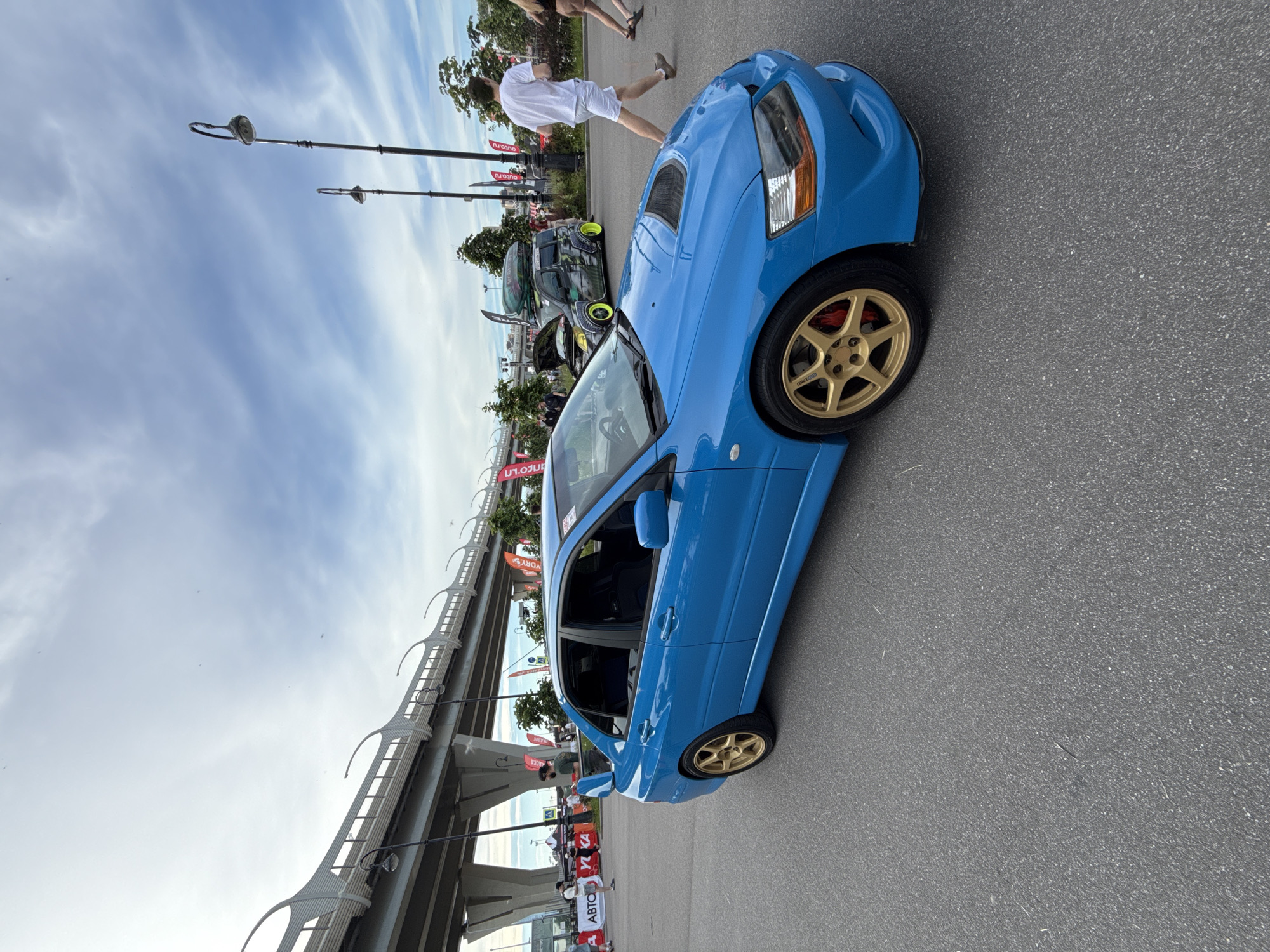 A blue Mitsubishi Lancer Evolution sedan is parked on an asphalt surface. The car has gold five-spoke wheels and a rear spoiler. In the background, there is a multi-level highway overpass, several lampposts, and a second car with a roof rack. A few people are visible in the background, and flags with Russian text are displayed. The sky is partly cloudy. The location is near the Southern Arc of the Western High-Speed Diameter in Saint Petersburg, Russia.