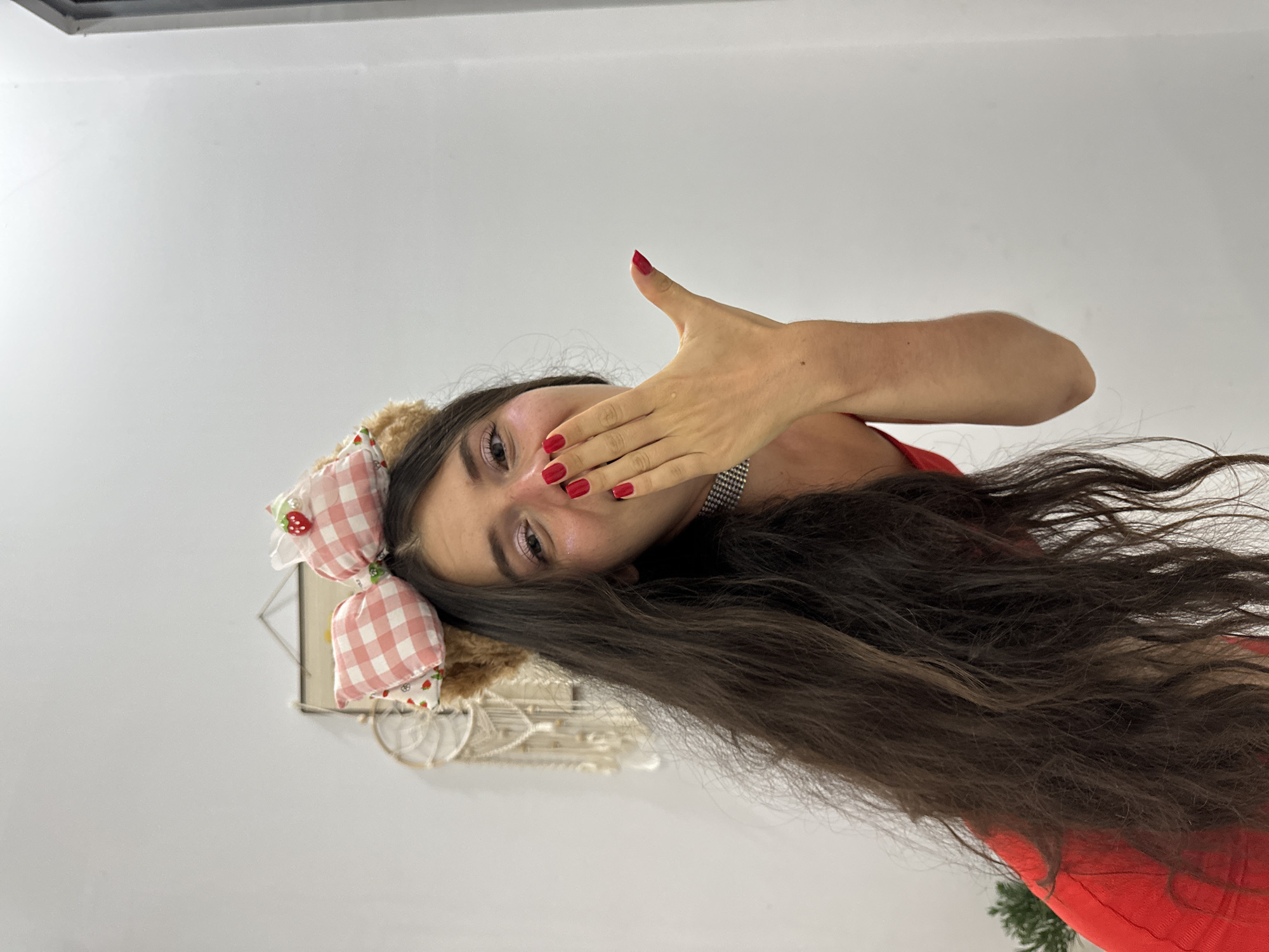 A woman with long, dark hair wears a headband with plush teddy bear ears and a pink gingham bow with a strawberry embellishment. She is dressed in a red top and a sparkly choker necklace. Her right hand is raised to her mouth, covering her lips as if blowing a kiss. Her fingernails are painted bright red. The background is a plain white wall with some decorative items hanging on it, including a macrame wall hanging. A hint of a green plant is visible on the left side of the frame. The lighting suggests an indoor setting. The image does not contain any information indicating its location in Mũi Né, Vietnam.