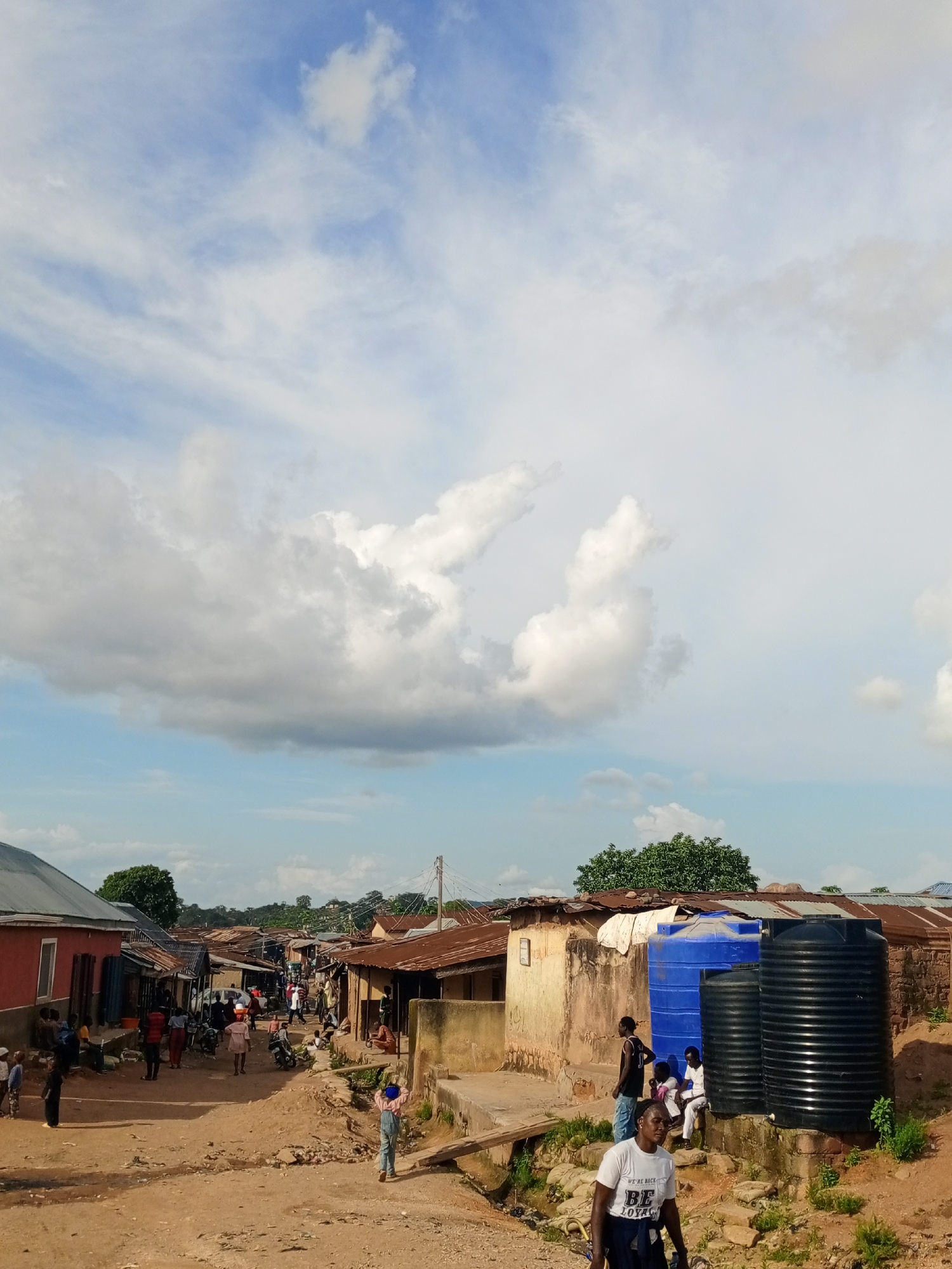 This is a medium shot image of a street in Jos, Nigeria, under a partly cloudy sky. The scene depicts a community with modest buildings made of corrugated metal and mud or cement. The road is unpaved and dusty. Several people are visible in the street, some walking, some sitting, and others engaged in conversation. A young boy in a blue shirt and overalls walks away from the camera down the street. A man in a white t-shirt with a logo on it is in the foreground, facing left. Two large water tanks, one blue and one black, are prominent in the middle ground, suggesting a communal water source. The buildings have a weathered appearance, and the overall impression is one of a lived-in, working-class neighborhood. There are several motorcycles parked along the street, indicating common modes of transportation. The presence of trees and vegetation suggests a somewhat rural or peri-urban setting within the city.