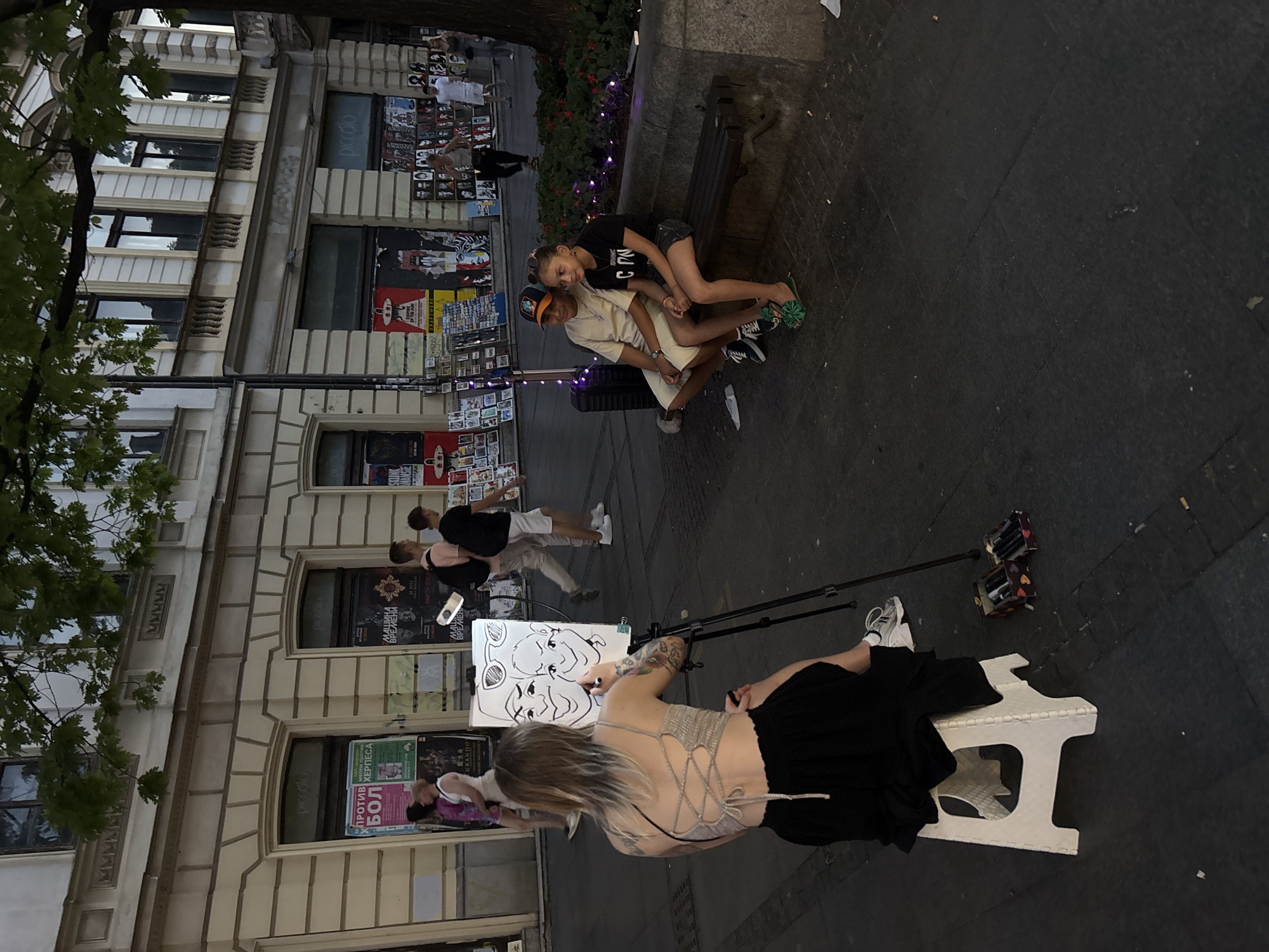 In Belgrade, Serbia, on a paved pedestrian walkway lined with shops and trees, a young woman with blonde hair, wearing a grey lace-up top and a black skirt, is seated on a white stool facing a tripod with a canvas. She is drawing a caricature of two individuals, a man and a woman. The man, seated on a stone bench to the right, is wearing a beige t-shirt and shorts. The woman, seated next to him, is wearing a black t-shirt and shorts. The bench is positioned in front of a raised flower bed. In the background, several people are walking, and various posters are displayed on the shop fronts. Two boxes containing markers are visible on the ground near the artist.