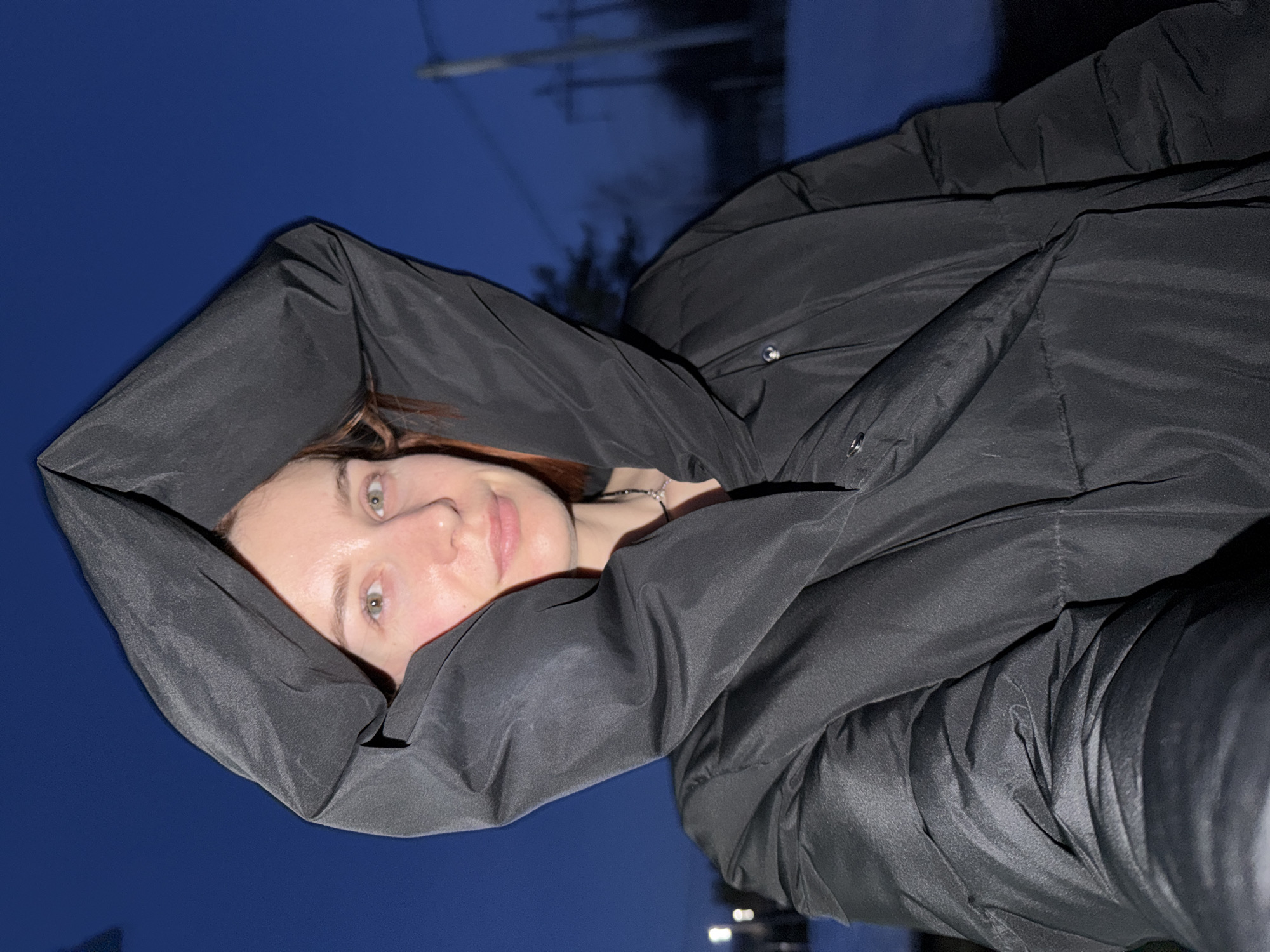 A woman with fair skin and red hair is looking at the camera. She is wearing a black puffer jacket with a hood, which is pulled up over her head. She also appears to be wearing a silver necklace. The background consists of a dark blue sky with some power lines in the distance. The location may be Chaikovsky, Russia, however there is nothing to confirm the location from this image.