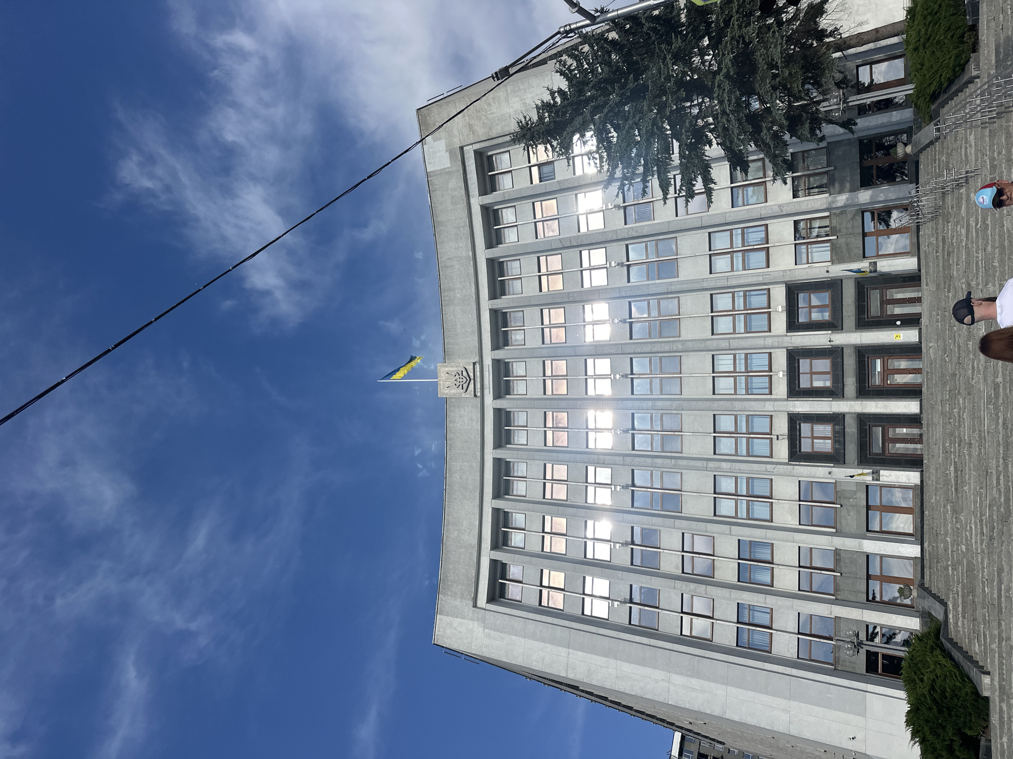 The image shows the Ternopil City Council building, a grand gray structure with many windows reflecting the bright blue sky and scattered white clouds. A Ukrainian flag flies proudly from the rooftop, accompanied by the national emblem. In front of the building, a large evergreen tree stands tall, its branches reaching towards the sky. Steps lead up to the entrance of the building, flanked by lush green bushes. At the bottom of the steps, two individuals are visible from behind, one wearing a black baseball cap and the other a blue and red cap. The sun casts a strong glare on the windows of the building, creating a bright, sunny atmosphere. The image is taken from a low angle, looking upwards at the imposing architecture.