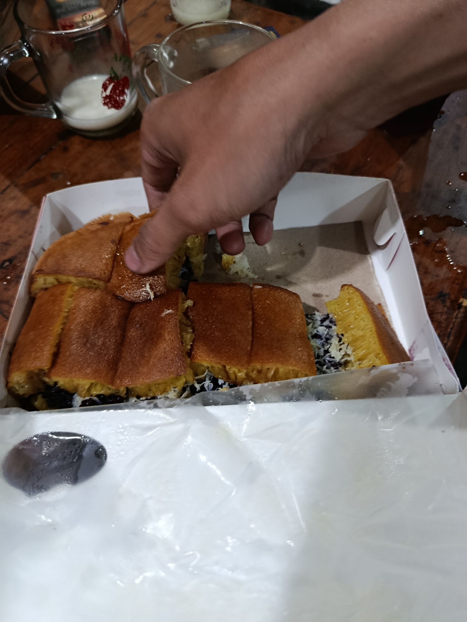 In this medium shot, a hand is reaching into a white cardboard box containing several pieces of sweet martabak, a popular Indonesian street food. The martabak is golden brown on the outside, with a fluffy, yellow interior. It appears to be filled with chocolate sprinkles and shredded cheese. The box is open, and the martabak is cut into rectangular pieces.
The scene is set on a wooden table, which is slightly wet in places, suggesting a recent spill or condensation. Two glass mugs, one with a red strawberry graphic, are visible in the background, both containing a white liquid, likely milk or a sweet beverage. The overall lighting is dim, creating a warm and intimate atmosphere, suggesting the image was taken indoors or in the evening. There is no visible text in the image. The activity depicted is someone reaching for a piece of martabak, possibly about to eat it. The context suggests a casual gathering or a moment of indulgence. The location is identified as Depok, Indonesia, which aligns with the presence of sweet martabak.