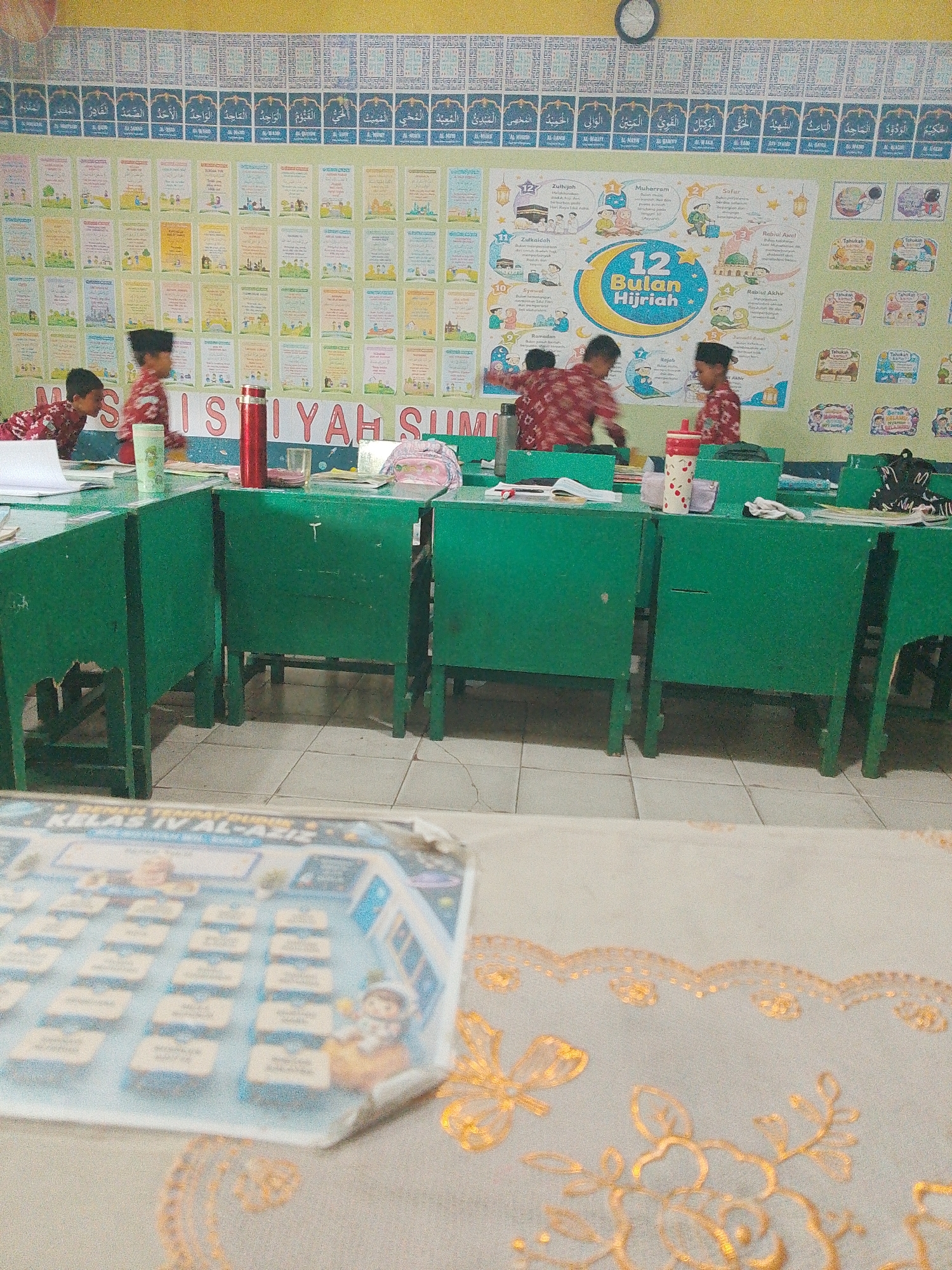 The image depicts the interior of a classroom, likely in Tembung, Indonesia, as indicated by the presence of Islamic educational materials and the overall aesthetic. The classroom is furnished with several green desks, some of which have visible textbooks and water bottles on them. In the background, a wall is adorned with numerous posters featuring Islamic calligraphy, illustrations related to Islamic months, and educational charts. The posters are arranged in rows and columns, creating a vibrant and informative display. Three children, wearing traditional Indonesian attire including batik shirts, are present in the scene. One child is seated at a desk, while two others are standing and appear to be interacting or engaged in an activity. The lighting suggests it is daytime, with natural light likely entering from an unseen window or source. The floor is tiled with light-colored squares. In the foreground, a blurred image of a book or educational material with an illustration of an astronaut can be seen. The scene conveys a typical learning environment in an Islamic educational institution.