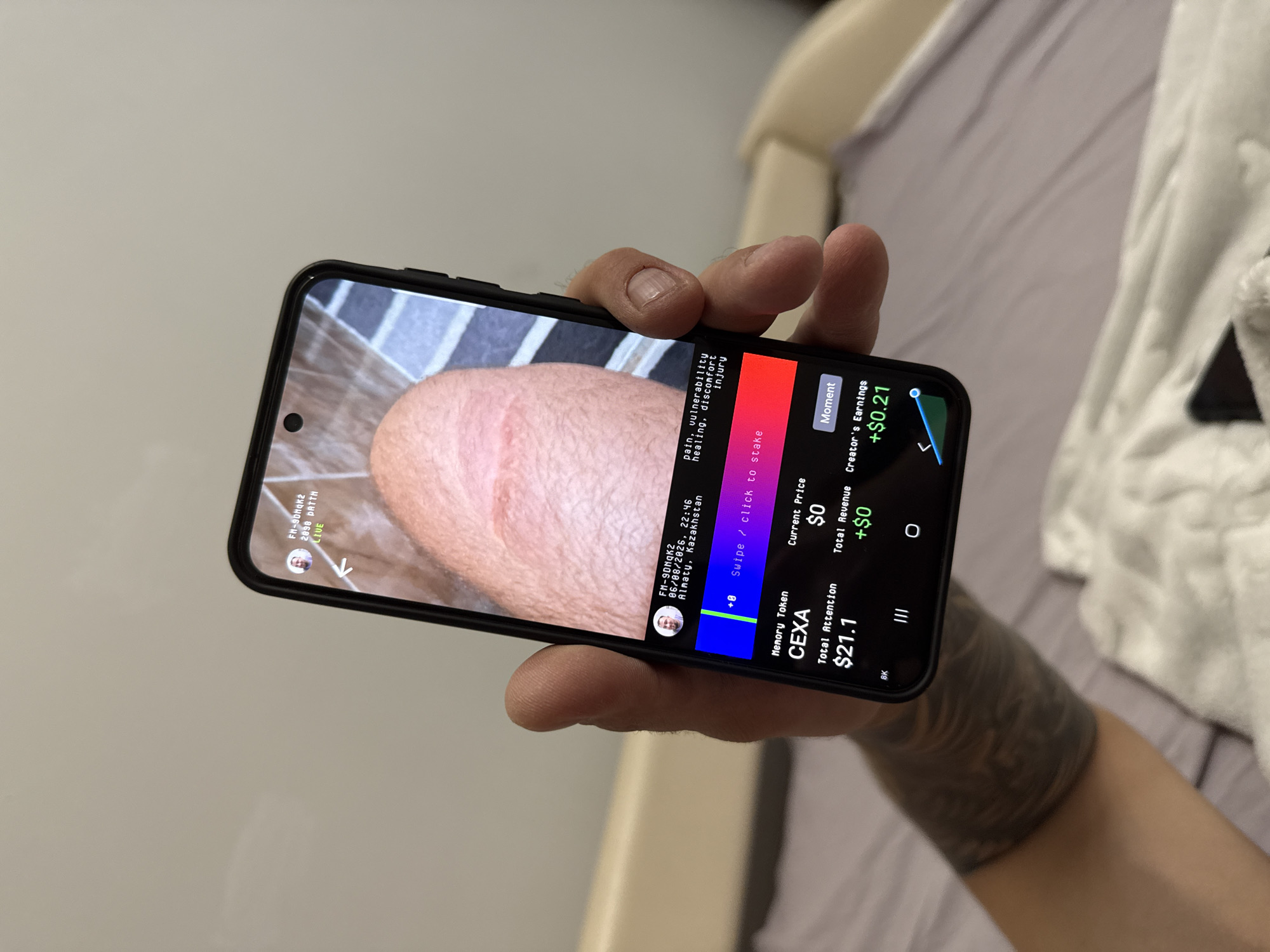 The image is a close-up, overhead shot of a hand holding a smartphone. The smartphone screen displays an application with various data points and a video feed. The video feed shows a close-up of a person's skin, possibly an arm or leg, with some redness and irritation visible. Text overlay on the screen indicates information such as "FM-9DHqk2", "2098 DATTN", and "LIVE", along with the location "Almaty, Kazakhstan", and a date "06/08/2026" at "22:46". Below the video feed, there are sections labeled "Memory Token CEXA", "Total Attention $21.1", "Current Price $0", "Total Revenue +$0", and "Creator's Earnings +$0.21". There is also a graphical representation of a line chart. The hand holding the phone has visible tattoos on the forearm. The background appears to be a bed with patterned bedding.