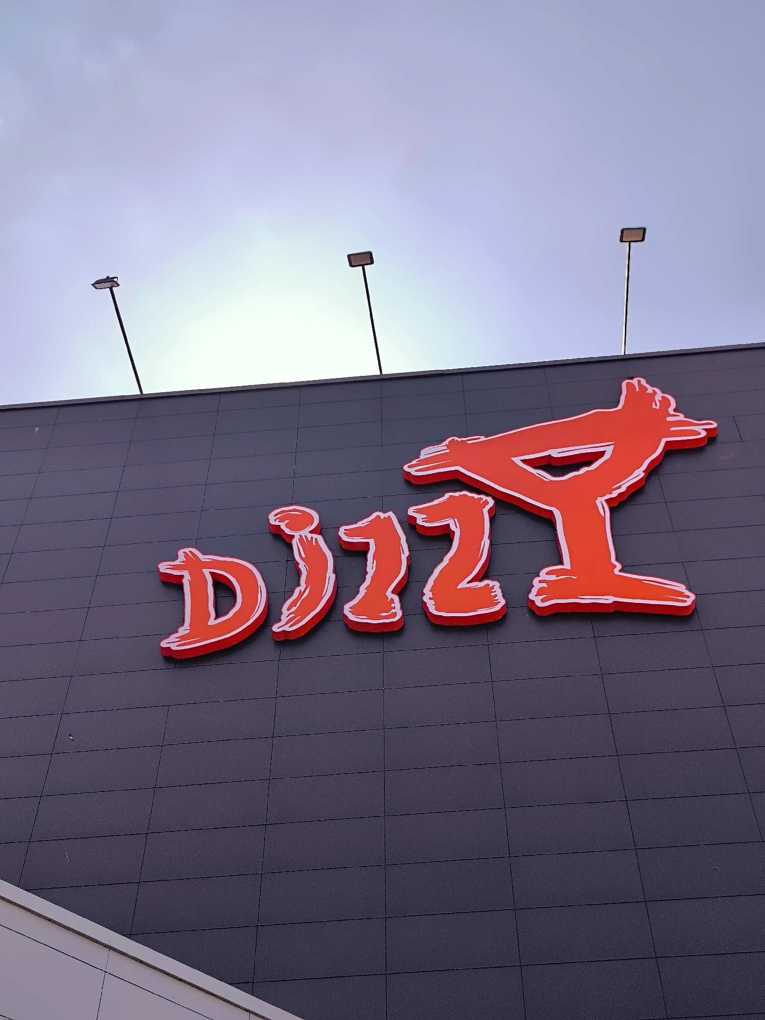 The image shows a close-up, low-angle shot of the red, illuminated "DIZZY" logo mounted on a dark gray, grid-patterned building facade. Two tall poles with rectangular lights are visible against a pale, overcast sky. The logo is stylized, with a white outline and a brushstroke effect. The building appears to be a commercial establishment. The context provided about Qarağandı city administration, Kazakhstan, suggests this is a sign for a business located within or near that administrative area.