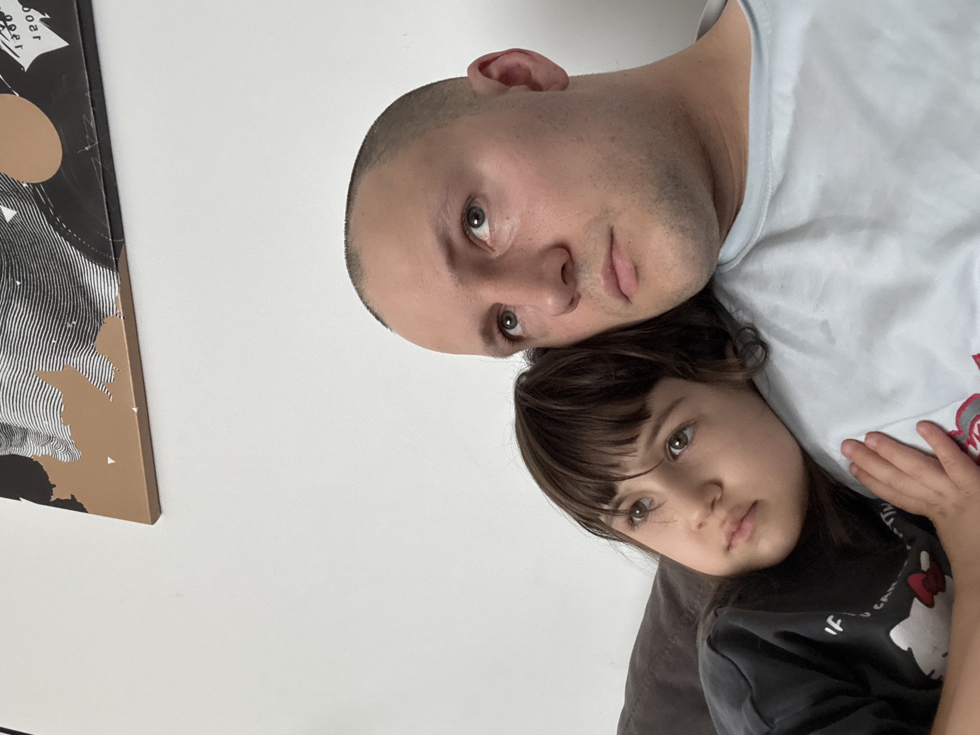 A man and a young girl are posed closely together. The man has short, light-colored hair and is wearing a light-colored t-shirt. The girl has brown hair and is wearing a dark-colored shirt with a print on it. A framed artwork hangs on a white wall. The location is Krasnoyarsk, Russia.