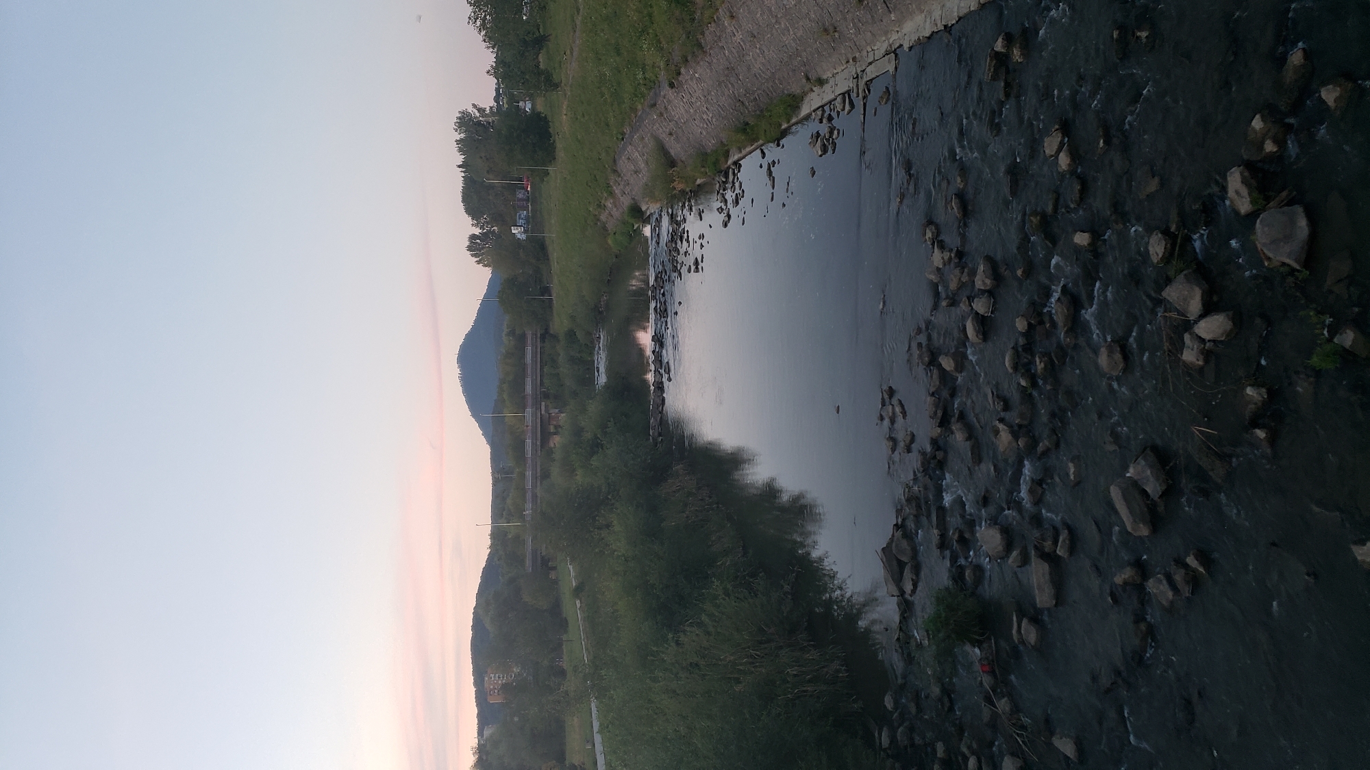 The image depicts a view of the Torysa River in Prešov, Slovakia, near the Sekčov district. The river flows from the foreground towards a bridge in the mid-ground. The water is relatively calm with some ripples, and it is dotted with numerous rocks, particularly in the foreground where they create a shallow, rocky bed. On the left bank of the river, there is dense, overgrown vegetation. The right bank is lined with a concrete embankment, with grass and trees beyond it. In the distance, a prominent conical hill, likely Šariš Castle hill, dominates the skyline, with a bridge spanning the river in front of it. The sky is a pale blue with hints of pink near the horizon, suggesting it is either dawn or dusk. A few trees and buildings are visible on the distant banks. There are no people or animals visible in the scene.