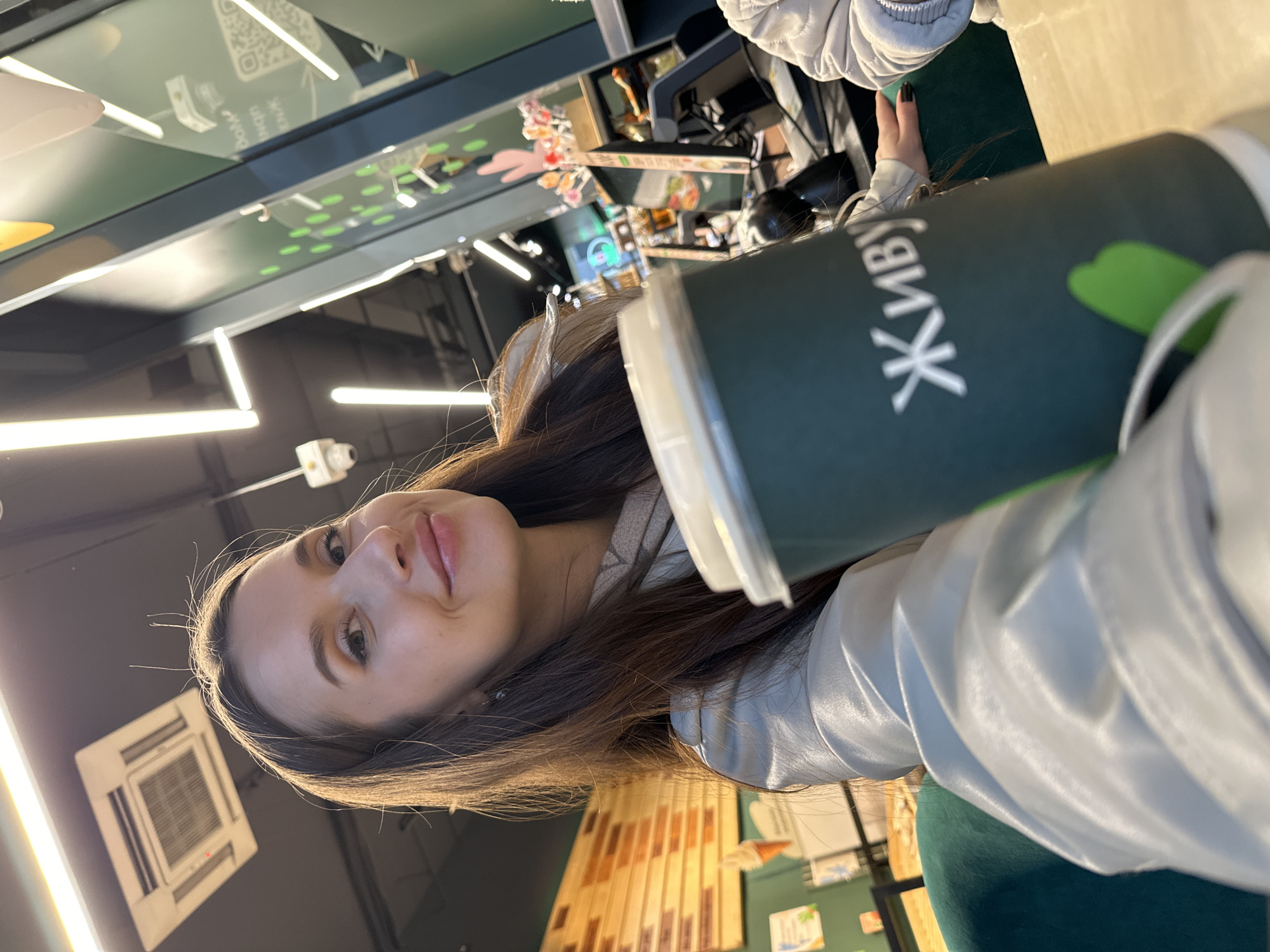 A young woman with long dark hair is captured in a selfie-style photograph, looking directly at the camera with a subtle smile. She is dressed in a light-colored, possibly white or silver, quilted jacket or coat. Her left hand is visible, holding a dark green disposable cup with a white lid, which is prominently featured in the foreground.
The most distinct visible text is on the cup itself, displaying the Cyrillic word "ЖИВУ" in white letters, which translates to "I LIVE" in Russian. This detail provides a strong location cue, aligning with the given context of Yekaterinburg, Russia.
The setting appears to be a modern indoor commercial space, likely a cafe, restaurant, or fast-food establishment. The background reveals dark walls or ceiling, bright linear LED lighting strips, and a white, rectangular air conditioning unit mounted on the ceiling to the left. Further in the background, there are hints of a service counter or display area, including reflections and what might be a screen with a QR code and some faint, unreadable Russian text. Wooden paneling can be seen on a wall in the mid-background to the right. The environment is well-lit by artificial lights, suggesting an indeterminate time of day. The woman appears relaxed and content.