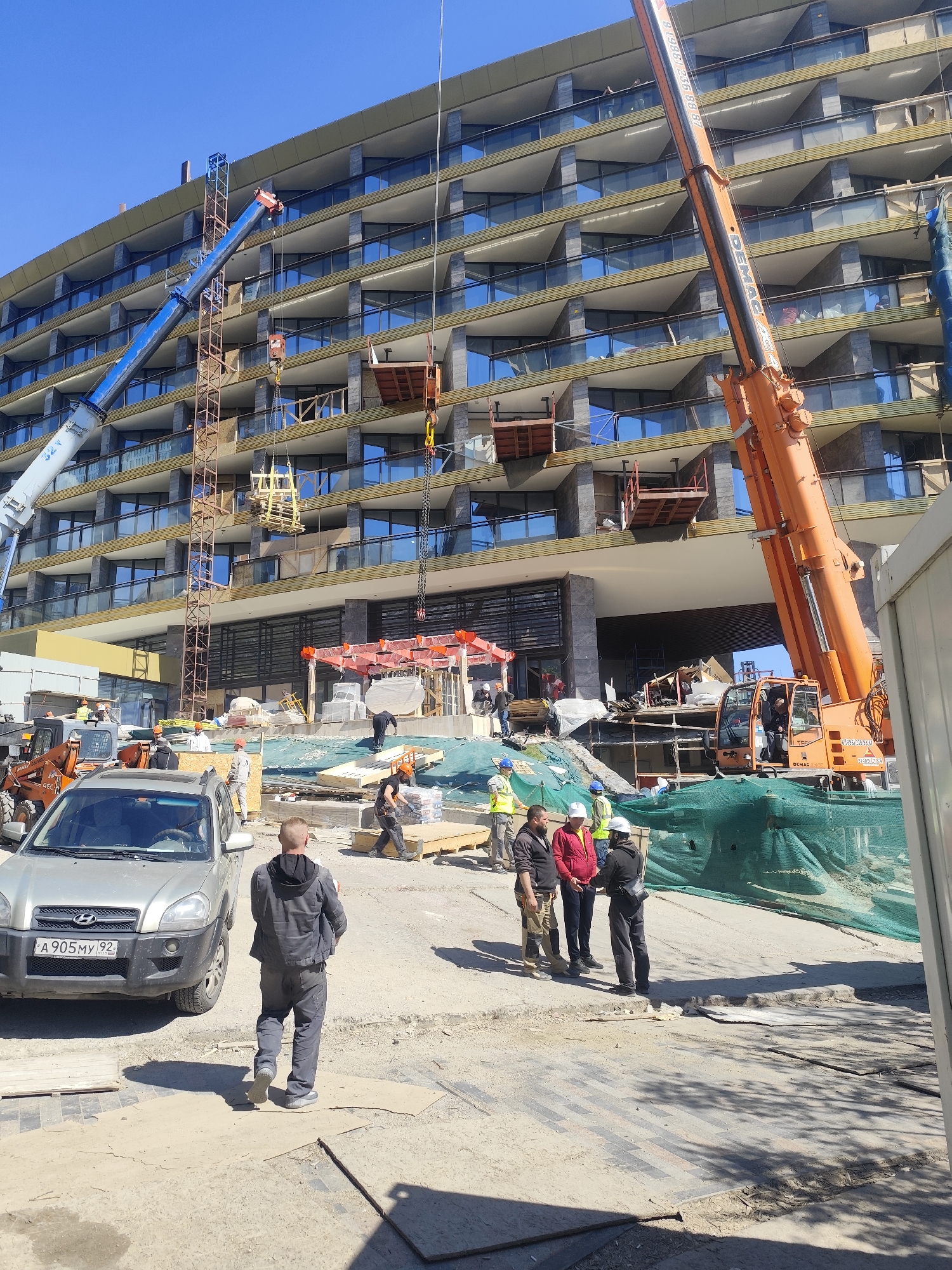 An active construction site is depicted on a bright, sunny day with a clear blue sky. A large, modern multi-story building, likely a hotel or residential complex, dominates the background, featuring numerous balconies with glass railings and stone paneling. The building appears to be in an advanced stage of construction or renovation, with some sections having exposed insulation or unfinished surfaces.
Two large cranes are prominent: a blue mobile crane on the left, extending its boom upwards, and an orange mobile crane on the right, also with its boom raised. The blue crane appears to be lifting a wooden frame structure, while the orange crane is positioned near the building's entrance. Various construction workers are scattered across the site. A group of several men, some wearing hard hats and safety vests, are gathered and conversing in the foreground to the right. Another worker, dressed in a dark jacket and pants, walks away from the camera towards a silver Hyundai Tucson SUV parked on the left. A yellow skid-steer loader is visible behind the SUV.
The ground is a mix of dirt and unfinished pavement, with wooden planks, construction materials, and green safety netting covering areas.
Visible text includes:
- On the blue crane: "32 Y".
- On the silver SUV's license plate: "A 905 MY 92".
- On the orange crane: "8 (988) 236 88 87" and "DEMAG" (twice).
- On the skid-steer loader: "460".
The license plate and phone number indicate the location is likely in Russia, with "92" on the plate typically corresponding to the Sevastopol region (Crimea) and "988" being a Russian mobile phone code.