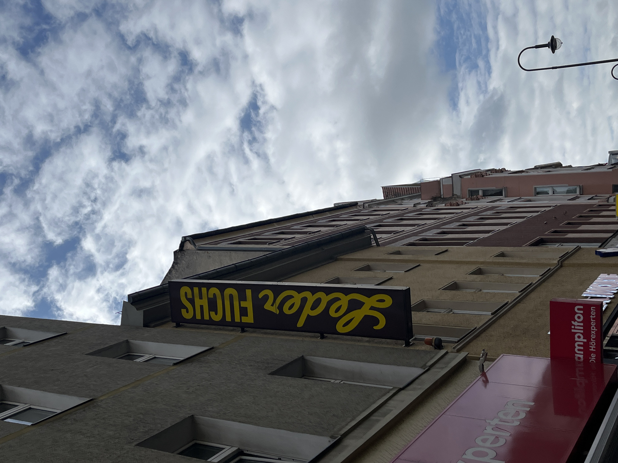 The image shows a vertical sign with yellow text on a dark background that reads "Leder Fuchs." The sign is attached to the side of a building, which appears to be in an urban environment. The building's facade is a neutral gray color with visible windows and trim. In the background, other buildings are visible, some with reddish-brown brickwork. The sky above is filled with clouds, suggesting an overcast or partly cloudy day. The overall scene depicts a street view in a city, likely a commercial area given the presence of the sign and other businesses visible in the background, such as one with a red sign and white text that reads "perten" and another red sign with white text that reads "amplifon Die Hörexperten". The image was taken from a low angle, looking upwards towards the buildings and the sky. The location is in Berlin, Germany.