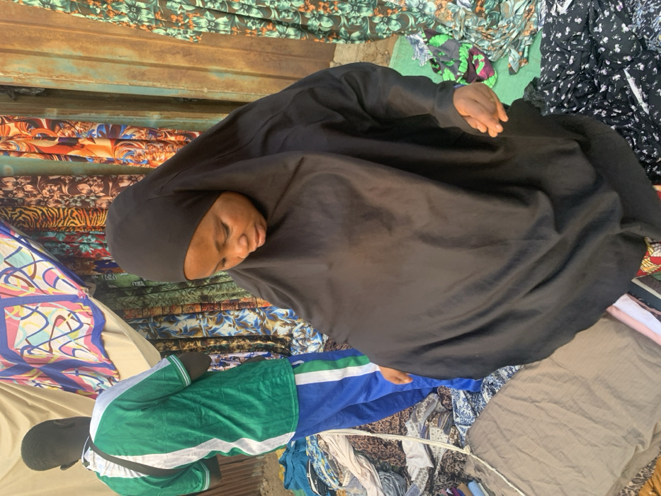 The image depicts an outdoor market scene in Okene, Nigeria, featuring two individuals amidst various fabric displays. In the foreground, a woman of African descent is seen from the chest up, wearing a plain black hijab covering her head and neck, and a loose-fitting black abaya-style garment. Her gaze is directed downwards and slightly to her right, and her right hand is visible near her hip, seemingly adjusting her clothing or reaching for an unseen object below. Behind her, to the left, a man is visible from the back, wearing a green and white striped short-sleeved shirt and blue athletic pants with a vertical green and white stripe.
The background consists of multiple fabric stalls. To the left, several rolls or lengths of colorful, patterned fabrics are draped and displayed, including designs with geometric shapes, animal prints, and floral motifs. A rusty, corrugated metal surface, likely part of a stall structure, is visible to the right of the fabrics. The ground beneath the individuals and stalls is unpaved, appearing as dirt or worn earth, and is scattered with various fabric scraps and debris, consistent with a bustling market environment. The scene is brightly illuminated by natural light.