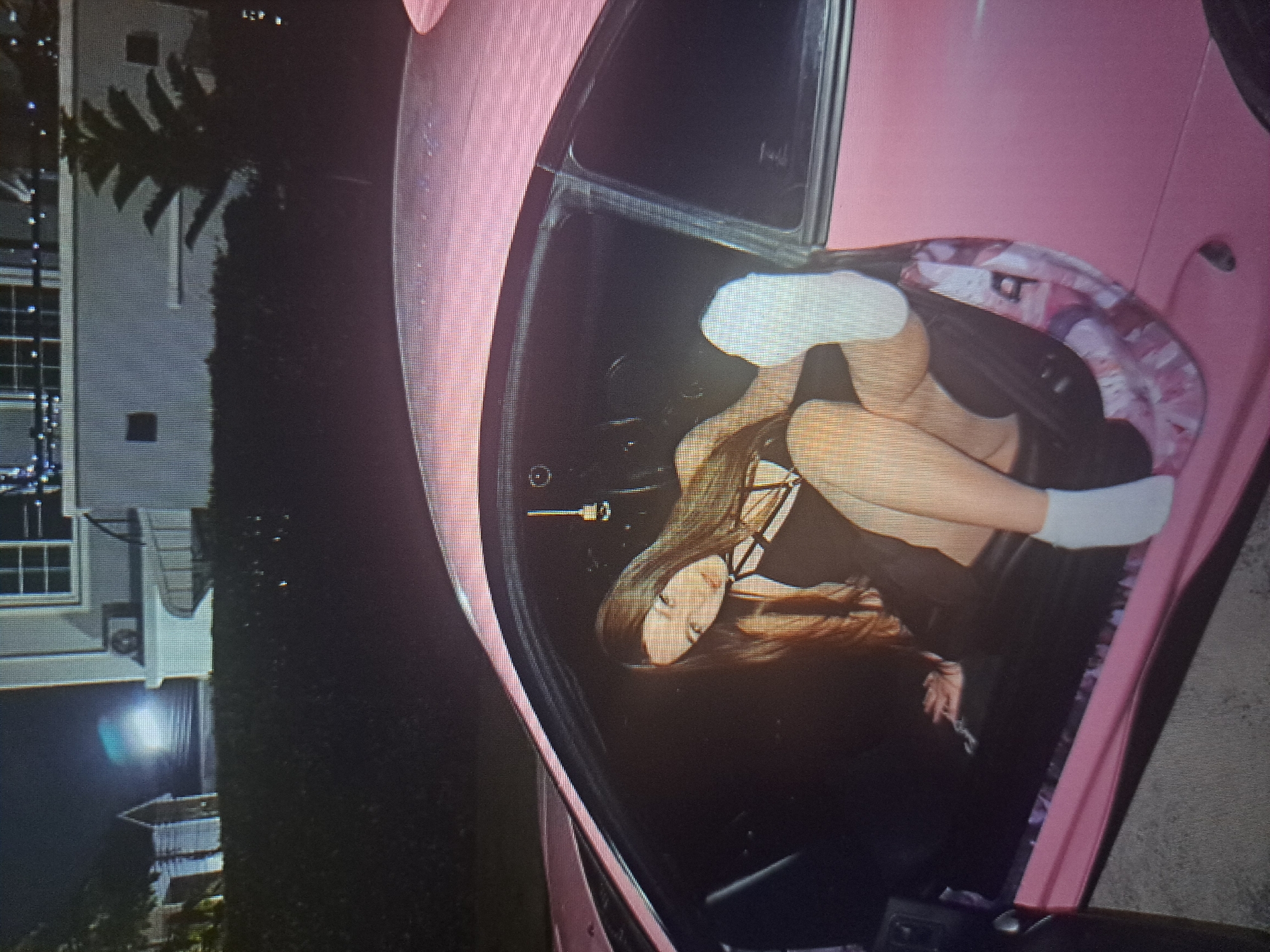 A pink car is parked on the side of a road at night, next to a house. The woman inside the car is curled up in the driver's seat. The specific location in Yerevan, Armenia cannot be determined from this image.