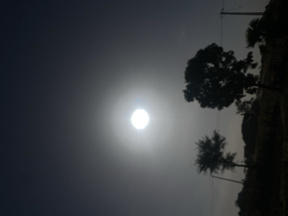 The image displays an outdoor, backlit scene in Rido, Nigeria. The sun, appearing as an overexposed, bright white orb with a prominent halo and a small blue lens flare, dominates the central upper portion of the frame. The sky transitions from bright white around the sun to a dark grey-blue gradient towards the edges. The lower half of the image consists of dark, silhouetted landscape features. These include two distinct trees in the midground: a smaller tree with a sparser canopy on the left, and a larger, denser-canopied tree to the right. A third, smaller, palm-like tree silhouette is visible on the far right. Two utility poles with visible wires are present: one on the left, partially obscured by the smaller tree, and another on the far right. The background features indistinct, mounded terrain or structures, all rendered in dark silhouette. No individuals are visible. The image possesses a soft focus throughout.