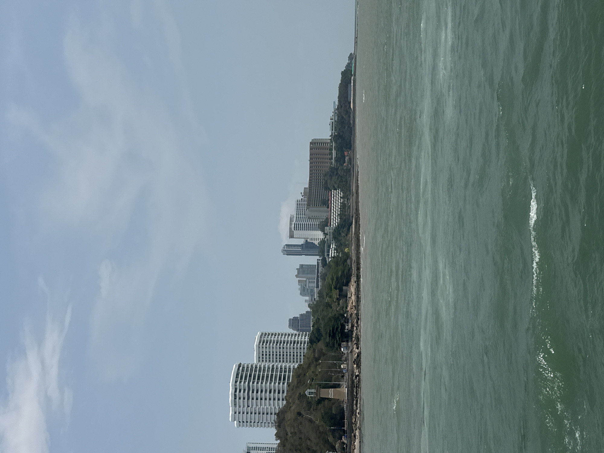 The image shows a coastal view of Pattaya City, Thailand. The scene depicts a skyline of buildings along the coast, with the sea stretching towards the right side of the frame. The sky is overcast, with pale clouds partially obscuring the blue. The water is a dark, murky green, with slight waves visible. The buildings vary in height and architectural style, including taller, modern structures and smaller, more traditional ones, interspersed with trees. The coastal strip has vegetation, contributing to a mix of urban and natural elements.
