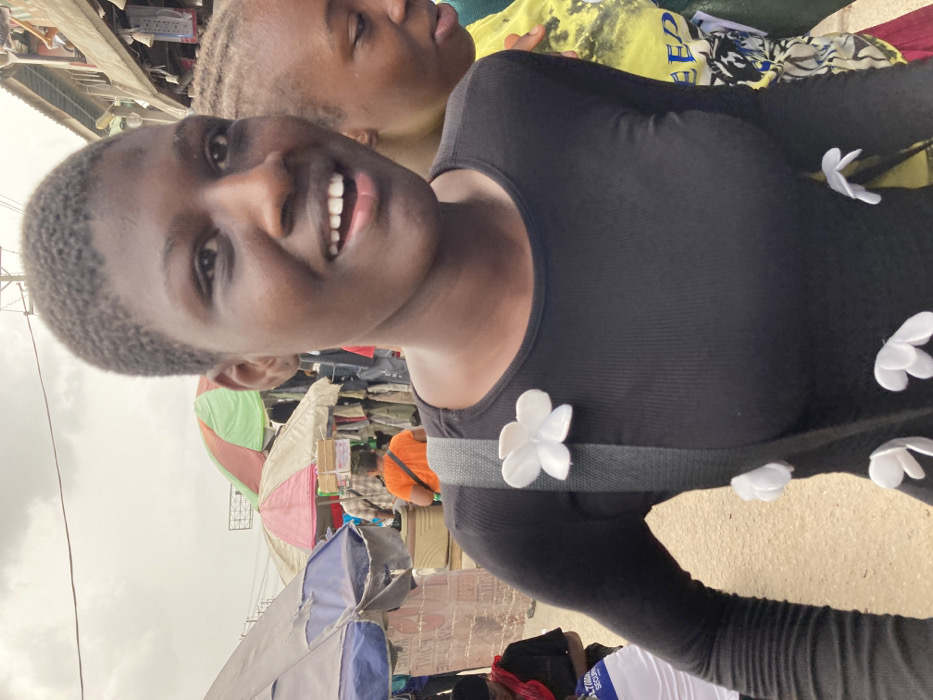 The image depicts a crowded outdoor market in Aba, Nigeria, under an overcast sky. In the foreground, a young Black woman with short, dark, closely-cropped hair smiles with her mouth open, revealing upper teeth. She wears a black, long-sleeved ribbed top embellished with multiple white, flower-shaped fabric appliques. A dark shoulder strap, also bearing a white flower applique, crosses her torso. To her immediate right, a second individual, partially obscured, shows braided hair and a yellow patterned shirt. The midground features market stalls beneath large, colorful umbrellas (blue-grey, red-white, green-brown), displaying hanging merchandise. An individual in an orange shirt is seen from behind, carrying stacked boxes on their head. Overhead power lines traverse the upper frame, and a building with a corrugated metal roof is visible on the far right. The ground appears as an unpaved surface.