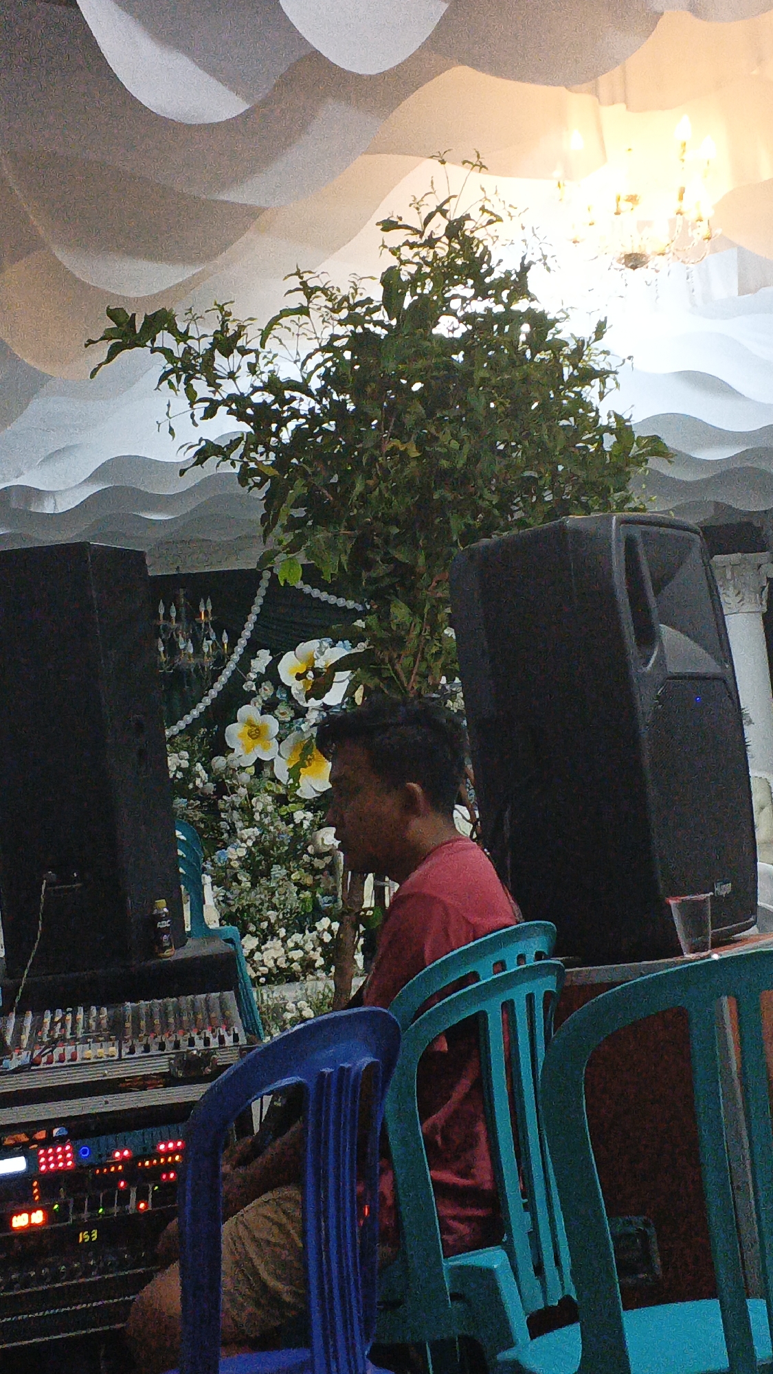 The image depicts a scene with a man sitting in front of a sound mixing board, in what appears to be an indoor event space. The man, wearing a red t-shirt, is partially obscured by two large black speakers positioned on either side of him. In the background, there is a decorative arrangement of artificial flowers and greenery, possibly for an event. Above, the ceiling is adorned with white, wavy fabric drapery, and a chandelier is visible. The overall impression is of a setup for a DJ or sound technician at a celebration or gathering, potentially in Soko, Indonesia, given the context provided. The lighting is warm, suggesting an ambient or decorative lighting scheme for the event. There are several plastic chairs in front of the sound board, one blue and others teal, some occupied by unseen individuals. A plastic cup is on a table to the right of the image.