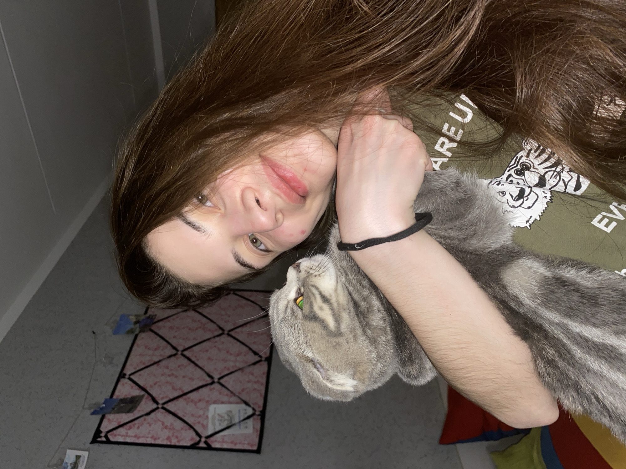 The image shows a young person with long, flowing brown hair, lying down and looking towards the camera with a gentle smile. Their face shows natural skin textures, including some minor blemishes or redness around the lips and on a cheek. They have light skin and brown eyes. The person is wearing a dark olive green t-shirt featuring a graphic of two stylized white and black tiger heads. Partial text is visible on the shirt, reading "ARE U" above the tiger graphic and "EVE" further down. A black elastic band or bracelet is worn on their left wrist.
Cuddled close to the person's arm and chest is a grey/silver tabby cat with prominent stripes and striking yellow-green eyes. The cat's face is clearly visible as it looks upwards, seemingly at the person. Both the person and the cat appear calm and comfortable, sharing a moment of affection.
The scene is indoors, likely a bedroom or living space, captured from an overhead and slightly tilted perspective. To the left, a plain grey wall is visible. The floor features a light-colored surface with a pink rug that has a black geometric grid pattern. Scattered on the floor near the rug are a few small papers or cards. In the bottom right corner, colorful fabric items, possibly cushions or blankets, are visible. The lighting is somewhat dim, suggesting an indoor environment, perhaps in the evening.