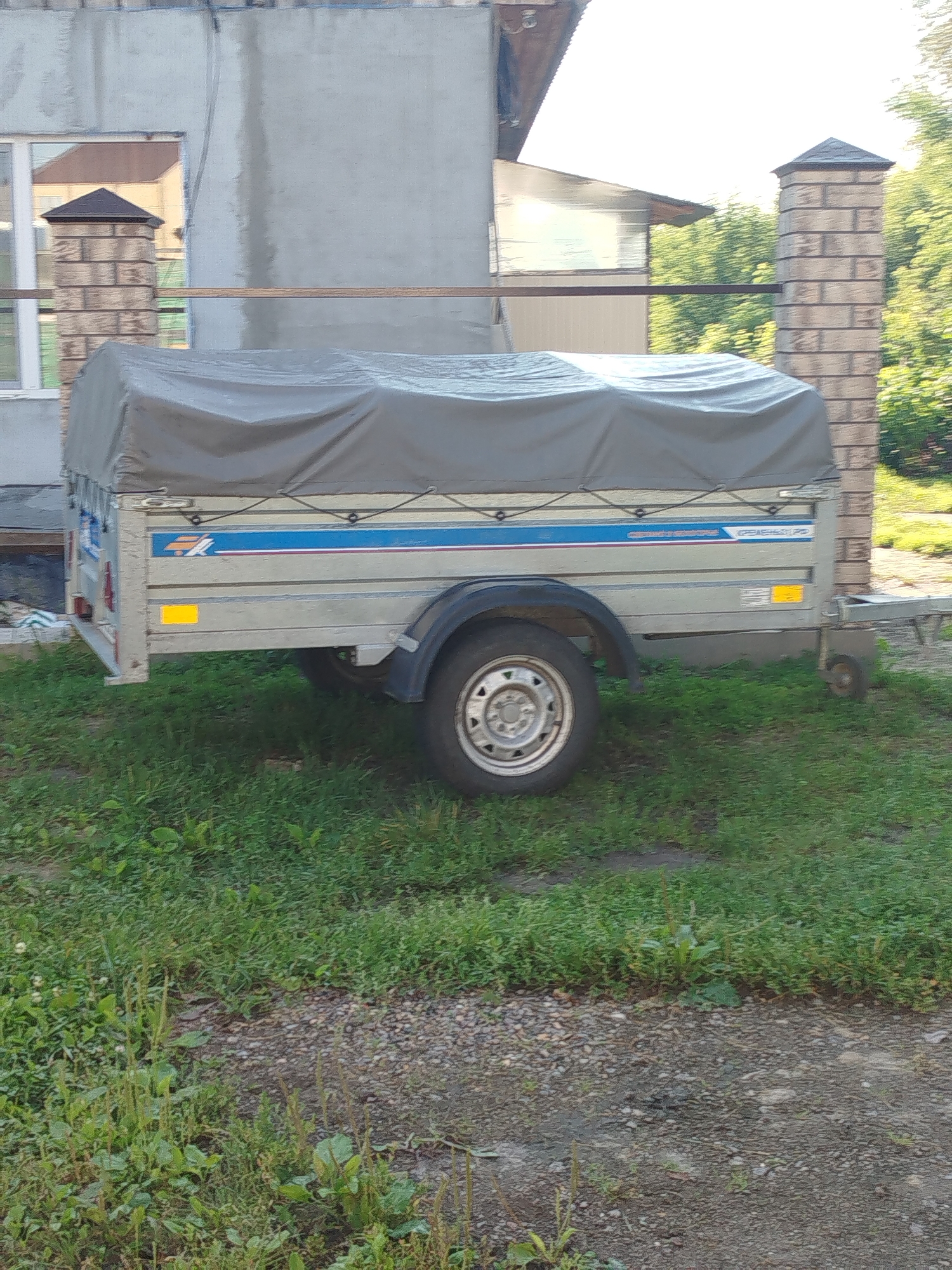 A silver trailer with a gray tarp cover is parked on a grassy area next to a gravel path. A brick pillar with a dark cap stands to the right of the trailer, and a building with a concrete wall and windows is visible behind it. The scene appears to be a residential or commercial property in Zavodoukovsk, Russia. There are no people or animals visible in the image.