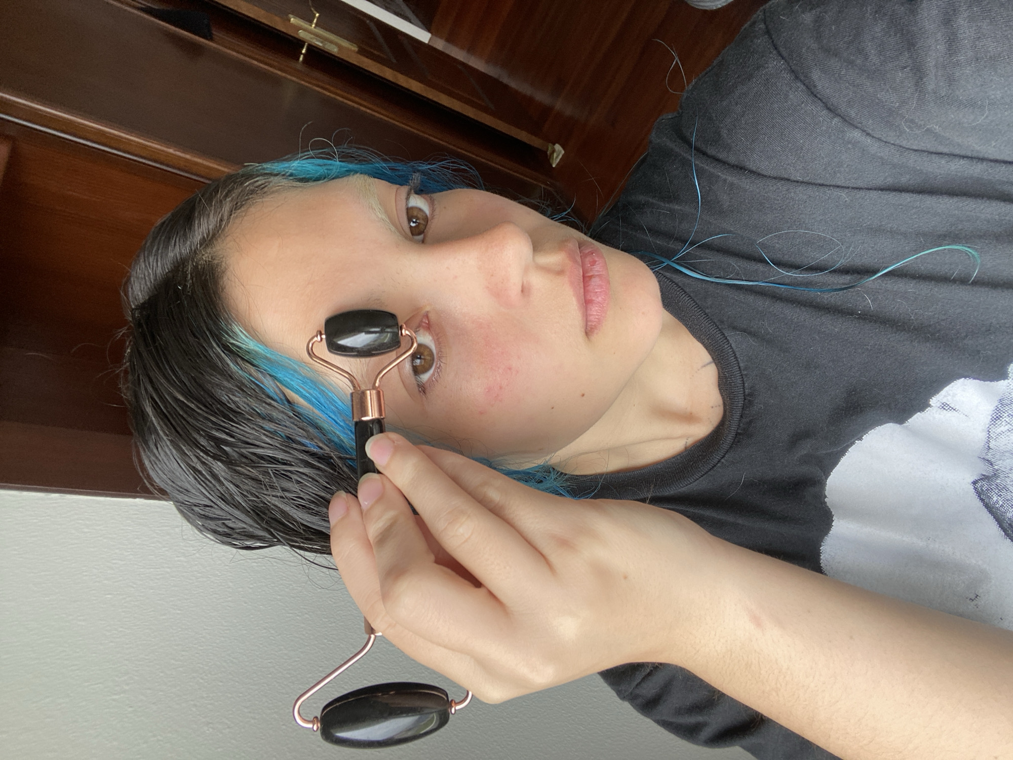 A woman is holding a facial roller to the right side of her face. The roller has a copper-colored frame and black stone rollers. The woman has black hair with blue highlights and is wearing a dark gray t-shirt. The background includes a wooden cabinet and a light-colored wall. The location is not specified and there is nothing present to indicate that it is in Gijón/Xixón, Spain.
