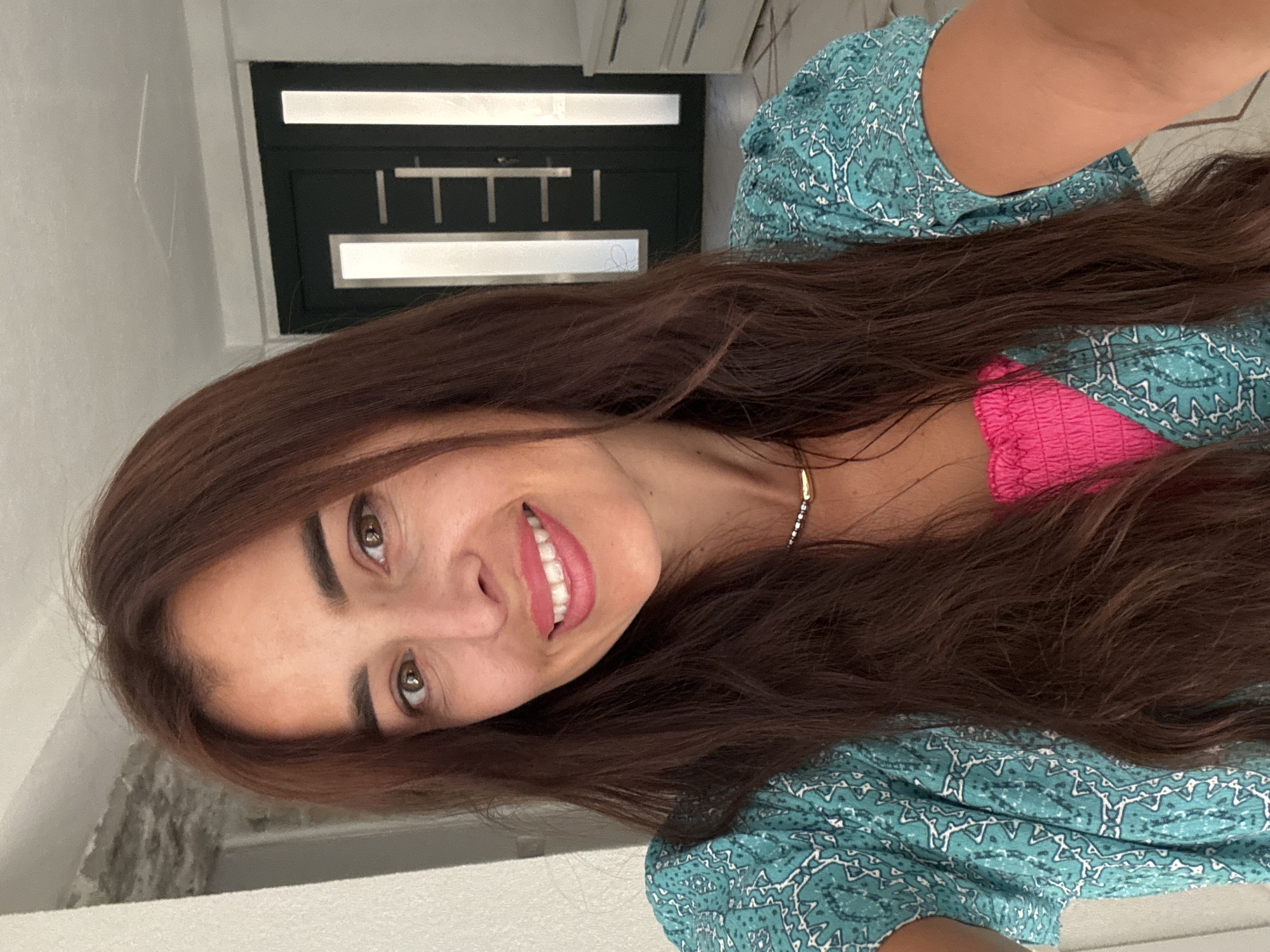 A woman with long, dark brown hair is taking a selfie. She is wearing a teal, patterned top over a pink strapless garment and a delicate necklace. She has brown eyes and is smiling, showing her teeth. The background appears to be an indoor setting with a dark-colored door with vertical glass panels visible behind her. To the right, a white dresser or cabinet is partially visible. A wall and part of a ceiling are also in the frame.