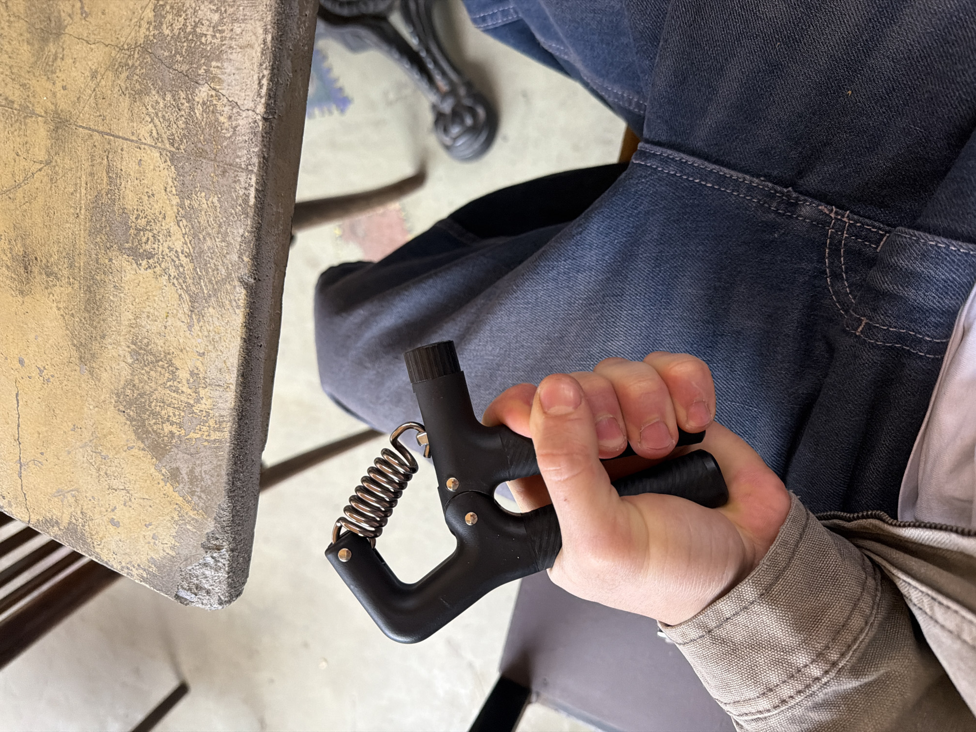 A person is seated, wearing blue jeans and a light-colored jacket. Their right hand holds a black hand grip strengthener. The scene is indoors, next to a weathered, light brown table with a chipped edge. The location is not discernible.