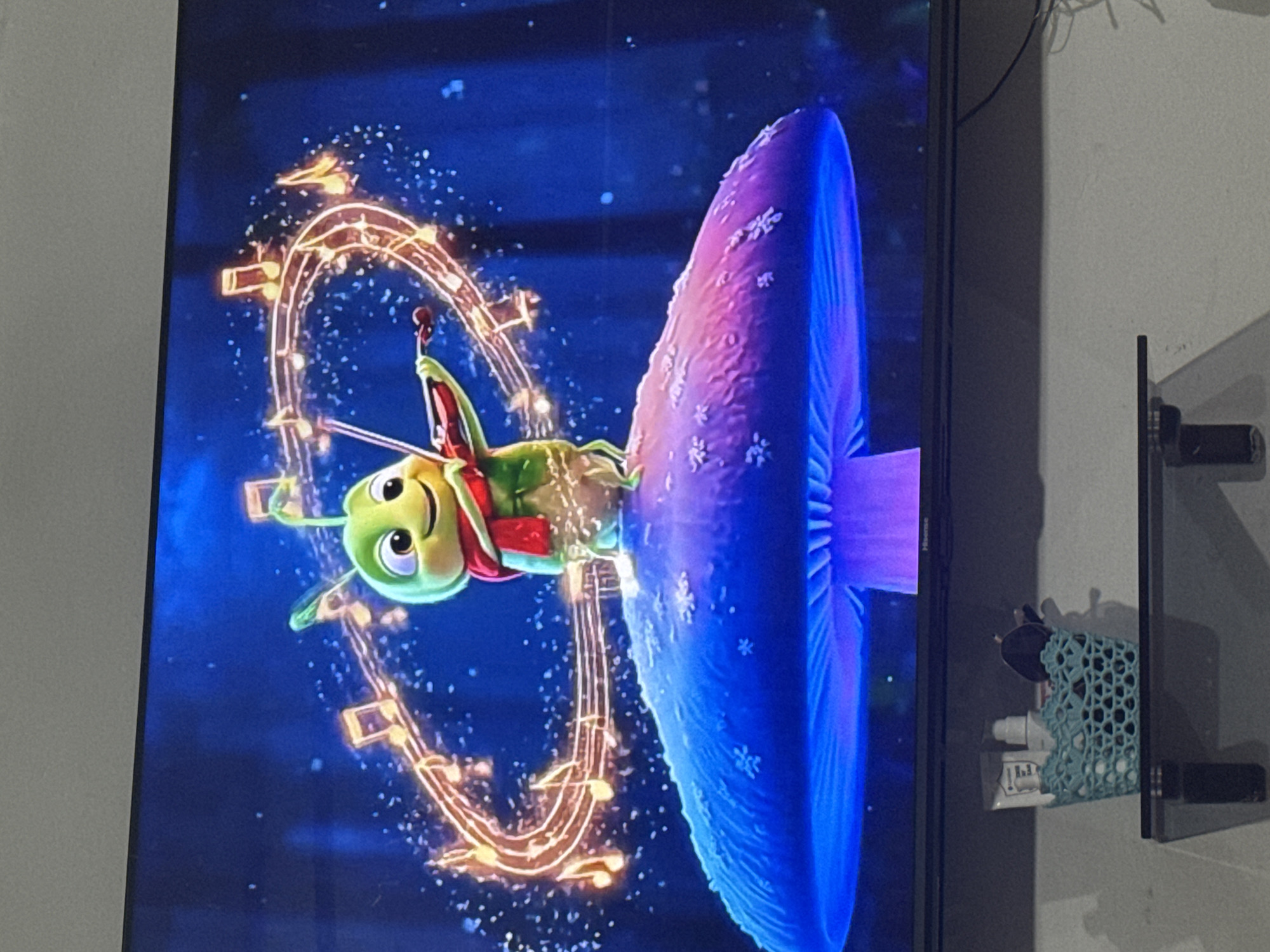 A television screen displays a cartoon image of a green grasshopper playing a violin while sitting on a large, bioluminescent mushroom. Musical notes are depicted in a glowing, circular pattern around the grasshopper. The background is a dark blue, starry night sky with faint trees. Below the television, a small blue basket containing toiletries and makeup brushes sits on a shelf. The provided location information, Chardara, Uzbekistan, is not visually evident in the image.
