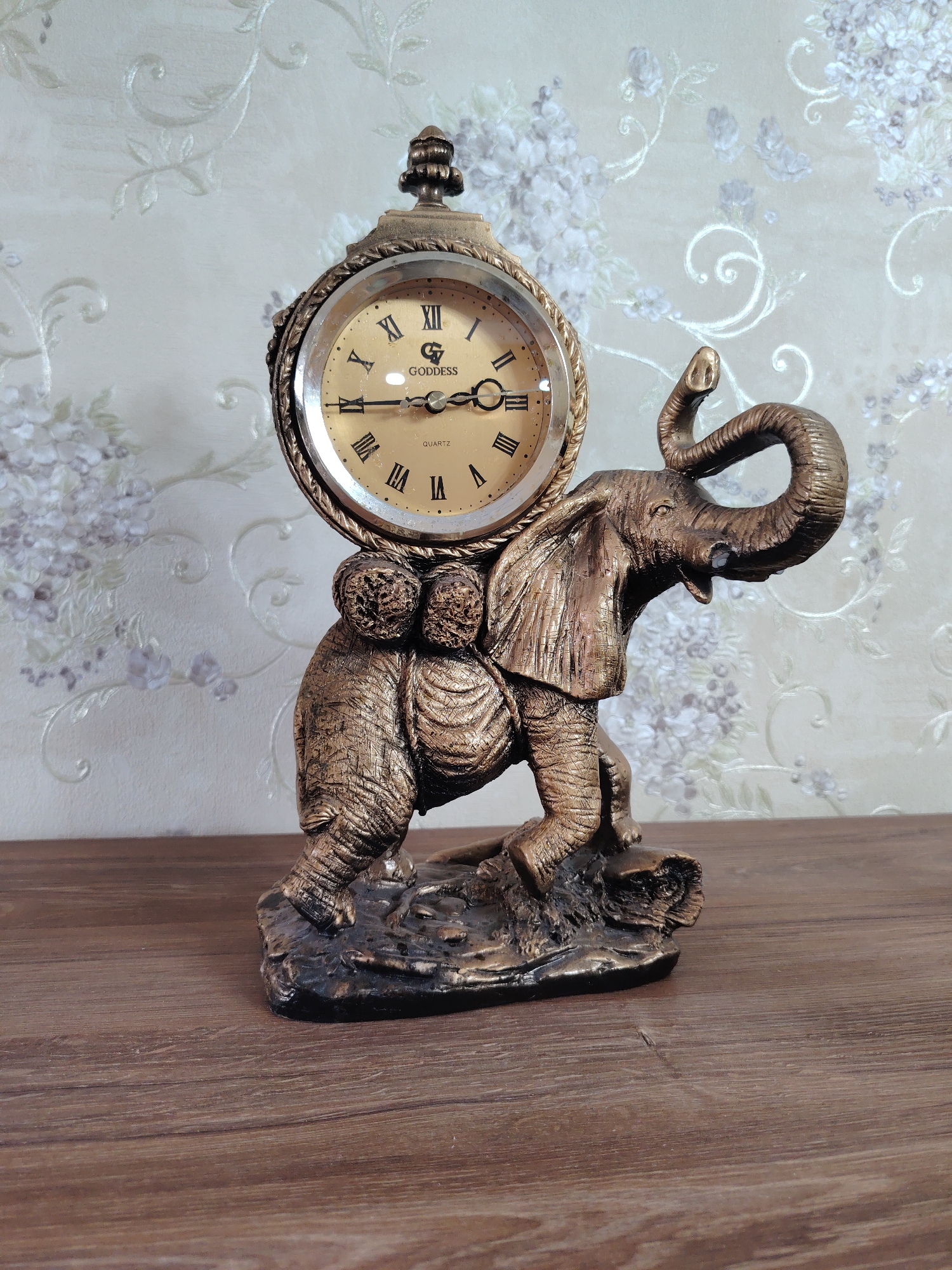 A close-up, eye-level, medium shot captures a decorative bronze-colored elephant figurine supporting a clock. The elephant, depicted in a dynamic pose with its trunk raised, stands on a dark, textured base. It is carrying what appear to be two large bundles on its back, securely attached. The clock face, positioned above the elephant, is round with a gold-colored dial. Roman numerals mark the hours, and the hands indicate a time slightly after 3:20. The word "GODDESS" is visible below the 12 o'clock position, and "QUARTZ" is printed near the 6 o'clock position. The background consists of a light-colored wallpaper with a subtle floral pattern in muted tones of grey and white. The figurine is placed on a wooden surface, likely a table, with a visible wood grain. The scene is well-lit, with soft shadows suggesting ambient lighting. The image is static and does not depict any active events or individuals. The photograph was taken indoors, likely in a domestic setting. The location is not specified in the image itself, and no information about Petropavl, Kazakhstan, is present within the visual data.