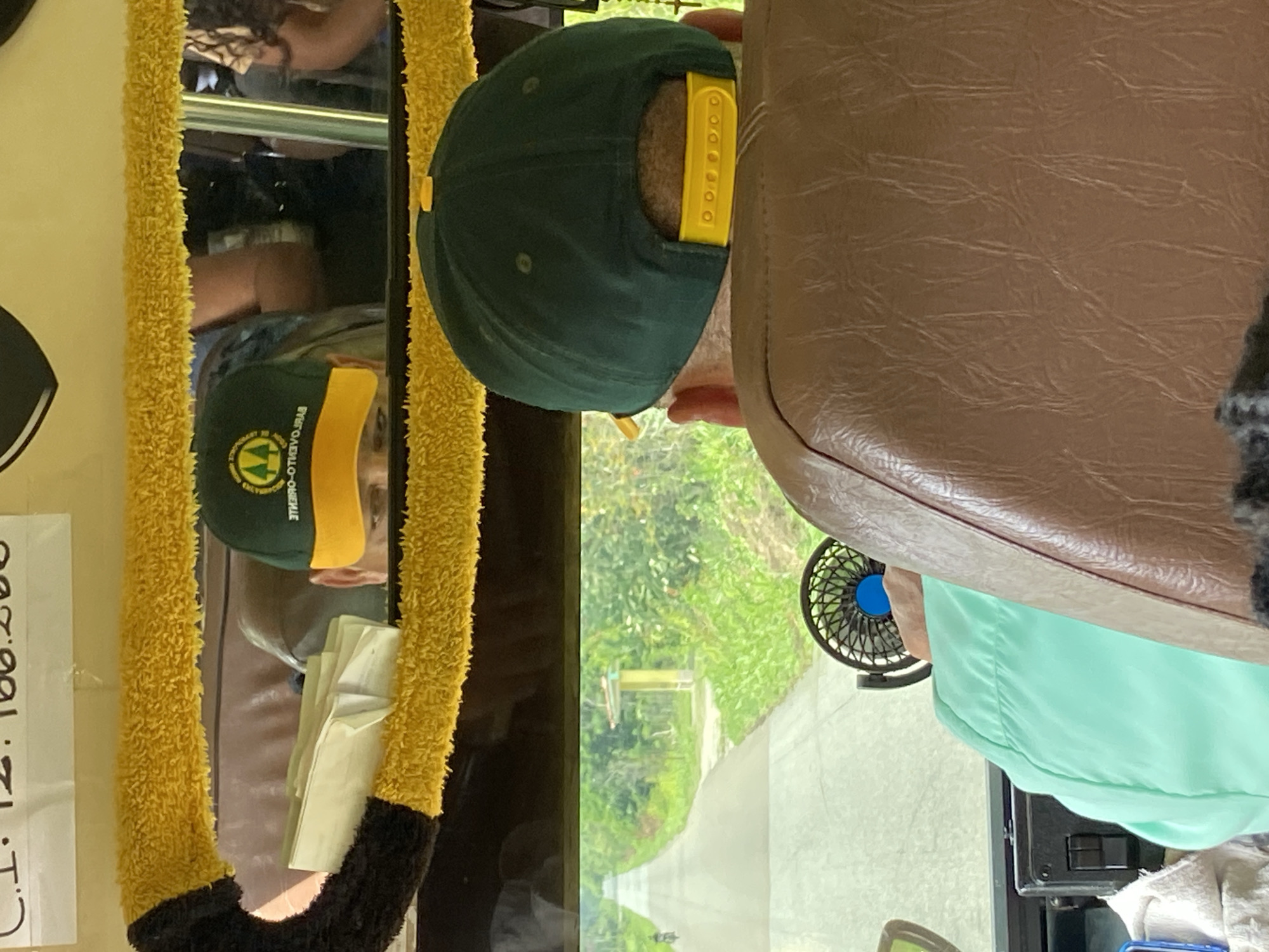 The image appears to be taken from inside a vehicle, looking forward through a windshield or window. In the foreground, the back of a person's head is visible, wearing a dark green baseball cap with a yellow brim and an emblem. The person is seated in a brown leather seat. To the left, a small black fan with a blue center is mounted.
The reflection in the mirror above shows another person wearing a similar dark green baseball cap with a yellow brim and an emblem. This person's face is partially visible, looking downwards. There are some papers or documents on a surface reflected in the mirror. A fluffy yellow and black trim borders the top of the mirror.
The environment outside the vehicle is a paved road stretching into the distance, lined with lush green vegetation. A motorcycle is visible on the road in the left distance. In the middle distance, there is a yellow pole structure with a blue sign on top, next to the road. The scene suggests a rural or semi-rural setting.
The text at the top of the image, partially visible, reads "C.1. 12.100.260". Based on the context of San José de Barlovento, Venezuela, this could be a registration number or identifier. The overall impression is a candid shot of individuals traveling in a vehicle, possibly a bus or van, in a tropical or subtropical environment.