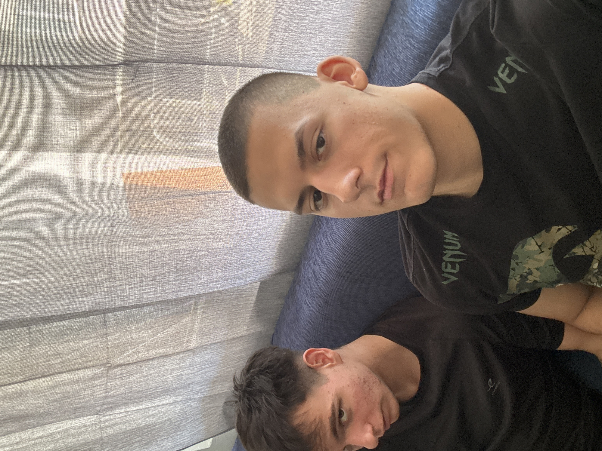 This portrait-oriented image captures two young men seated indoors in Plovdiv, Bulgaria. The perspective is tilted, showing the subjects angled from the bottom right towards the top left of the frame.
The foreground features a young man with a very short, dark buzz cut hairstyle. He has light skin with a few small blemishes or freckles visible on his forehead and cheeks. He looks directly towards the camera with a neutral to slightly thoughtful expression. He is wearing a black t-shirt with the brand name "VENUM" prominently displayed in green lettering on his right shoulder. The shirt also incorporates a subtle camouflage pattern.
Behind him, partially obscured and positioned lower in the frame, is another young man. His head and upper torso are visible. He has short, dark hair, styled with a slight fade on the sides. He is also wearing a black t-shirt, which has a small, indistinct white symbol or logo on the chest near the collar. His face is turned slightly away from the camera, showing a neutral expression.
Both men are seated on a blue, textured sofa or couch. The background is dominated by a light-colored, possibly grey, textured fabric, resembling curtains or blinds, with distinct vertical lines and some horizontal folds. Daylight appears to be filtering through this fabric, suggesting the photo was taken during the daytime. The setting indicates a casual, indoor environment.