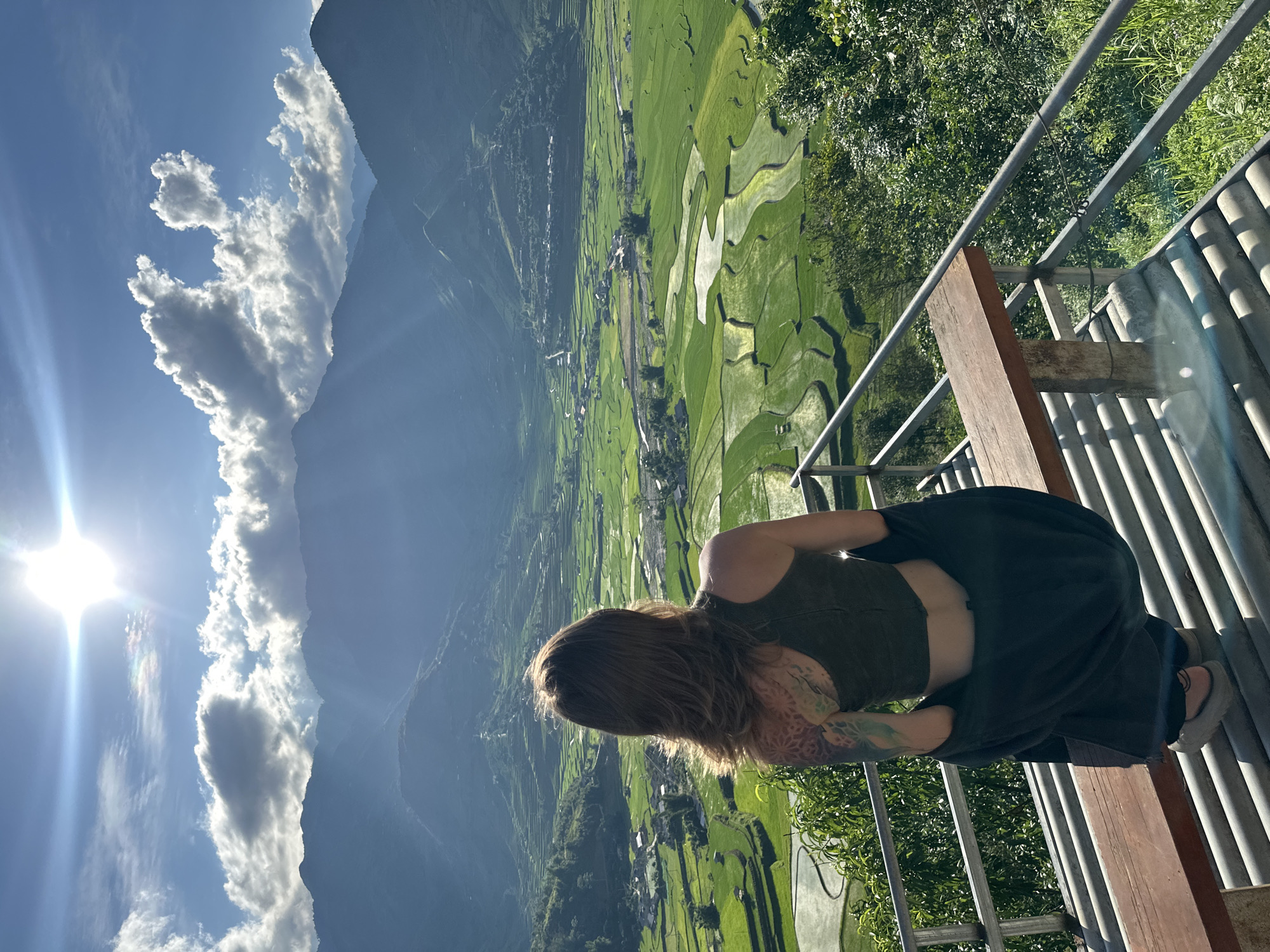 A female, viewed from behind, is seated on a wooden bench part of a viewing platform overlooking a vast agricultural valley in Tú Lệ, Vietnam. She has shoulder-length brown hair and wears a dark green crop top with a dark jacket draped around her waist. A colorful, intricate tattoo covers her left shoulder and upper arm. The platform features metal railings and a corrugated roof structure beneath the bench.
The expansive valley below is dominated by vibrant green terraced rice paddies, some fields containing water that reflects the sky. Small villages with houses are interspersed throughout the fields, and a winding body of water, likely a river or stream, is visible. In the background, majestic, hazy blue-grey mountains frame the valley under a bright blue sky with scattered white clouds. A prominent sun flare emanates from the upper left, casting strong light across the scene, and a faint rainbow-like optical phenomenon is visible near the sun.